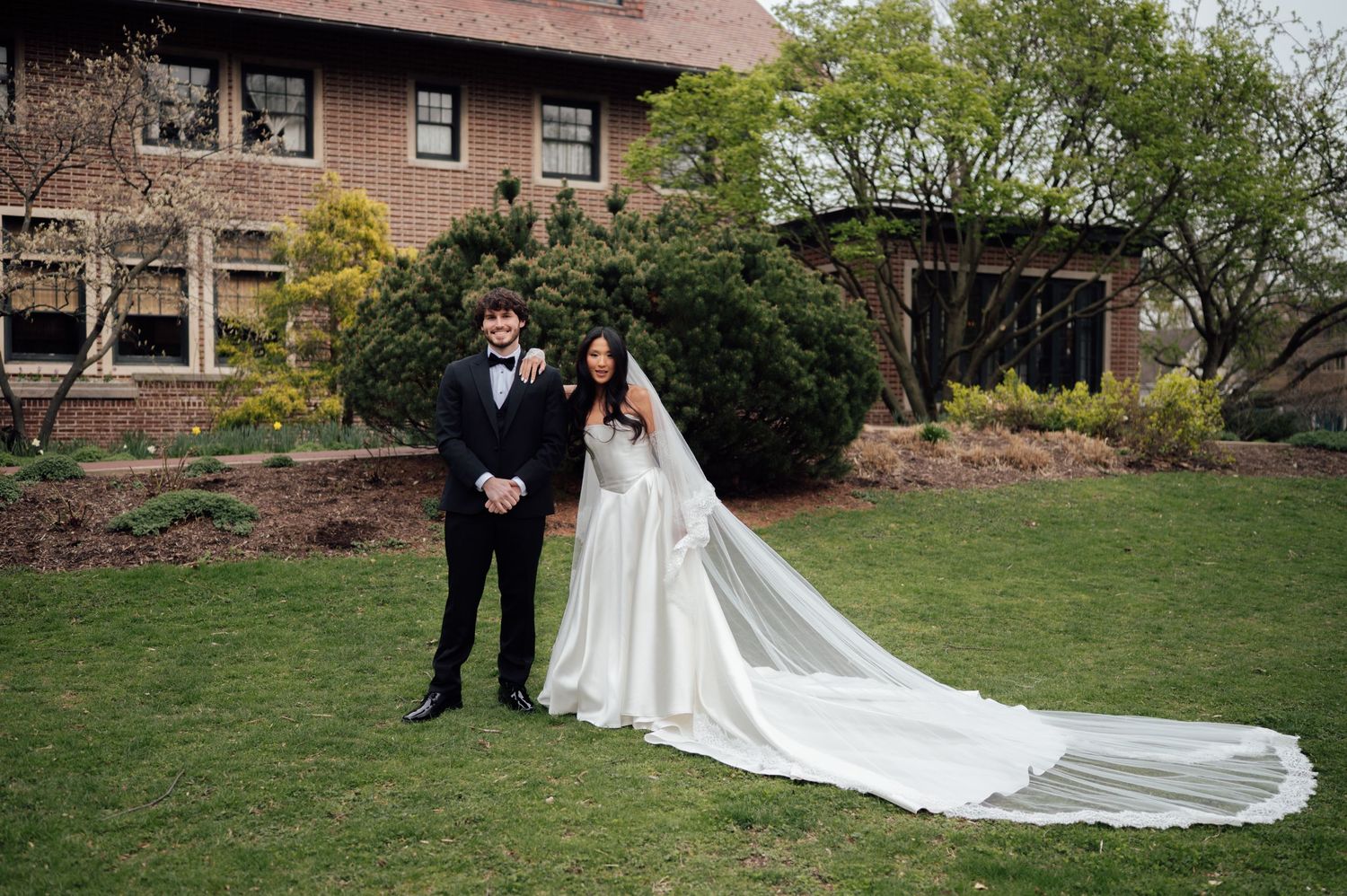 Bride in flowing white gown with long veil stands beside groom in black tuxedo on green lawn outside brick building.