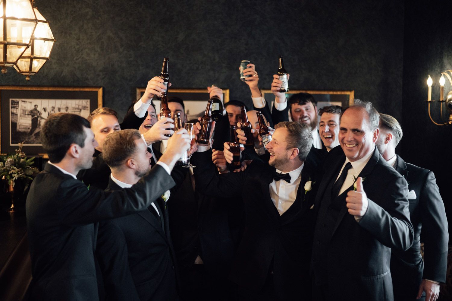 Groomsmen in black tuxedos raising drinks in celebratory toast at wedding reception.