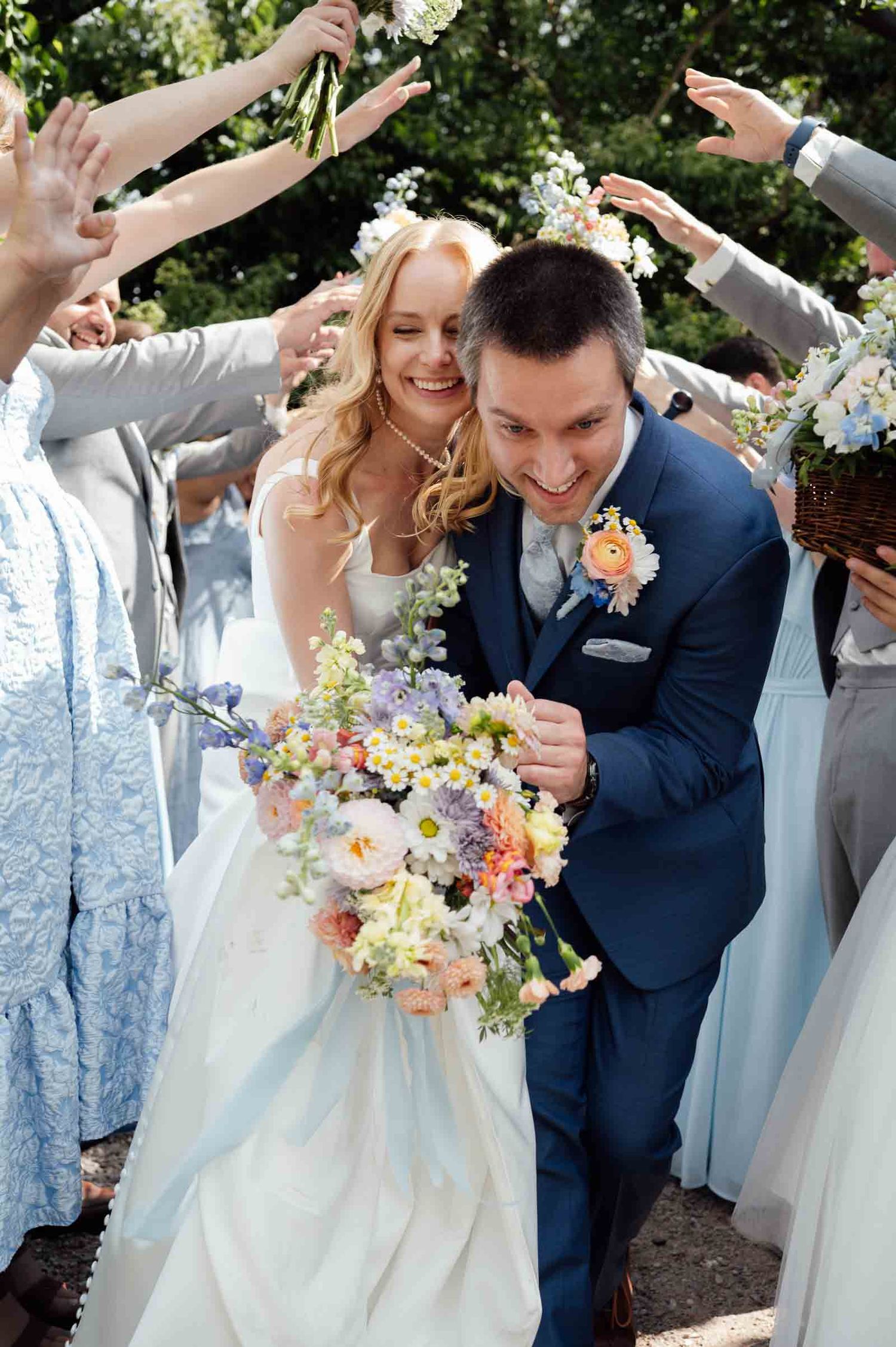 Joyful newlyweds walk through flower-tossing tunnel of guests after outdoor garden wedding ceremony.