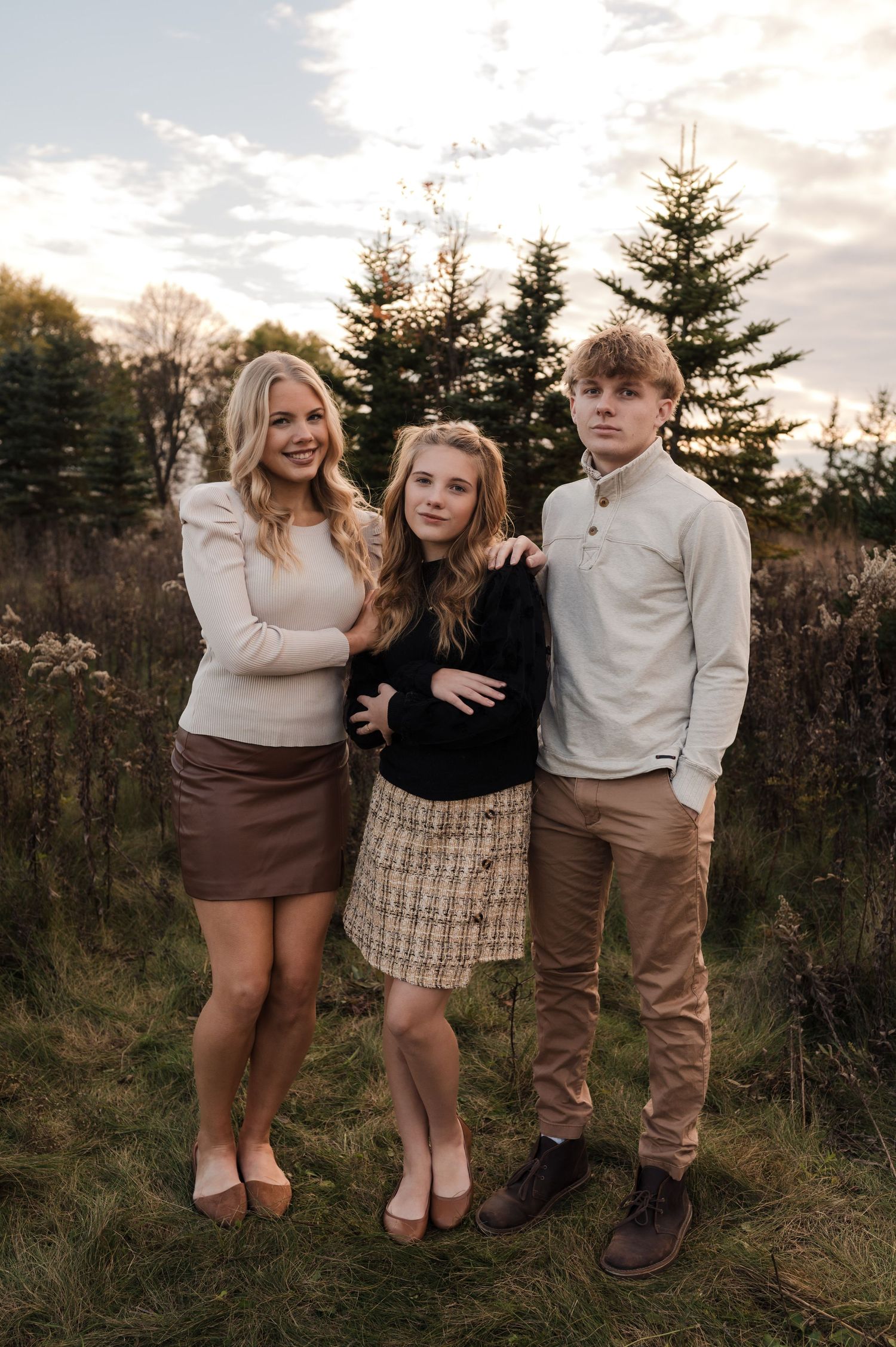 Three siblings smiling together outdoors at golden hour near pine trees in coordinated neutral outfits.