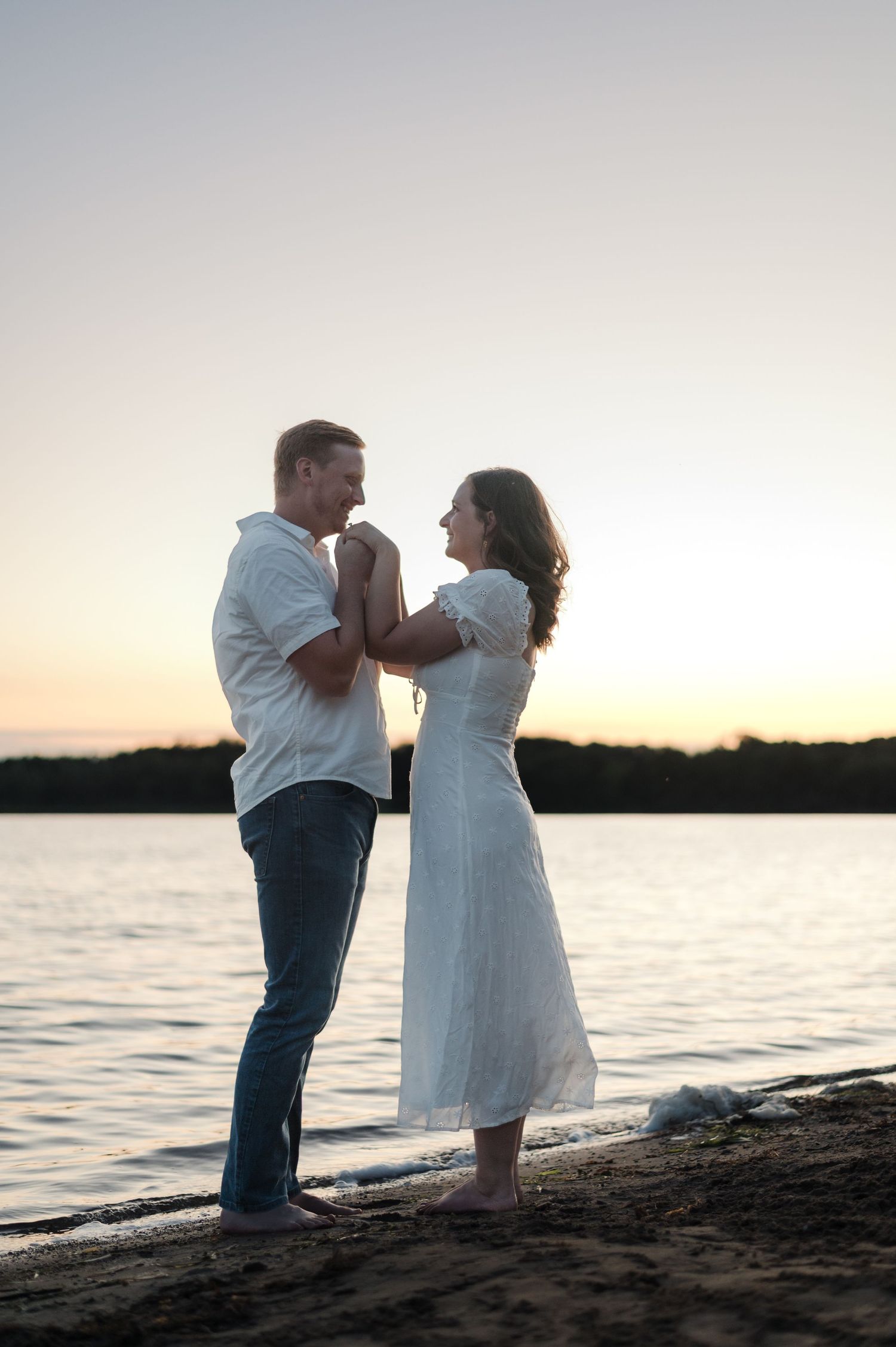 Engaged couple holds hands and smiles at each other barefoot on a sandy beach at sunset.