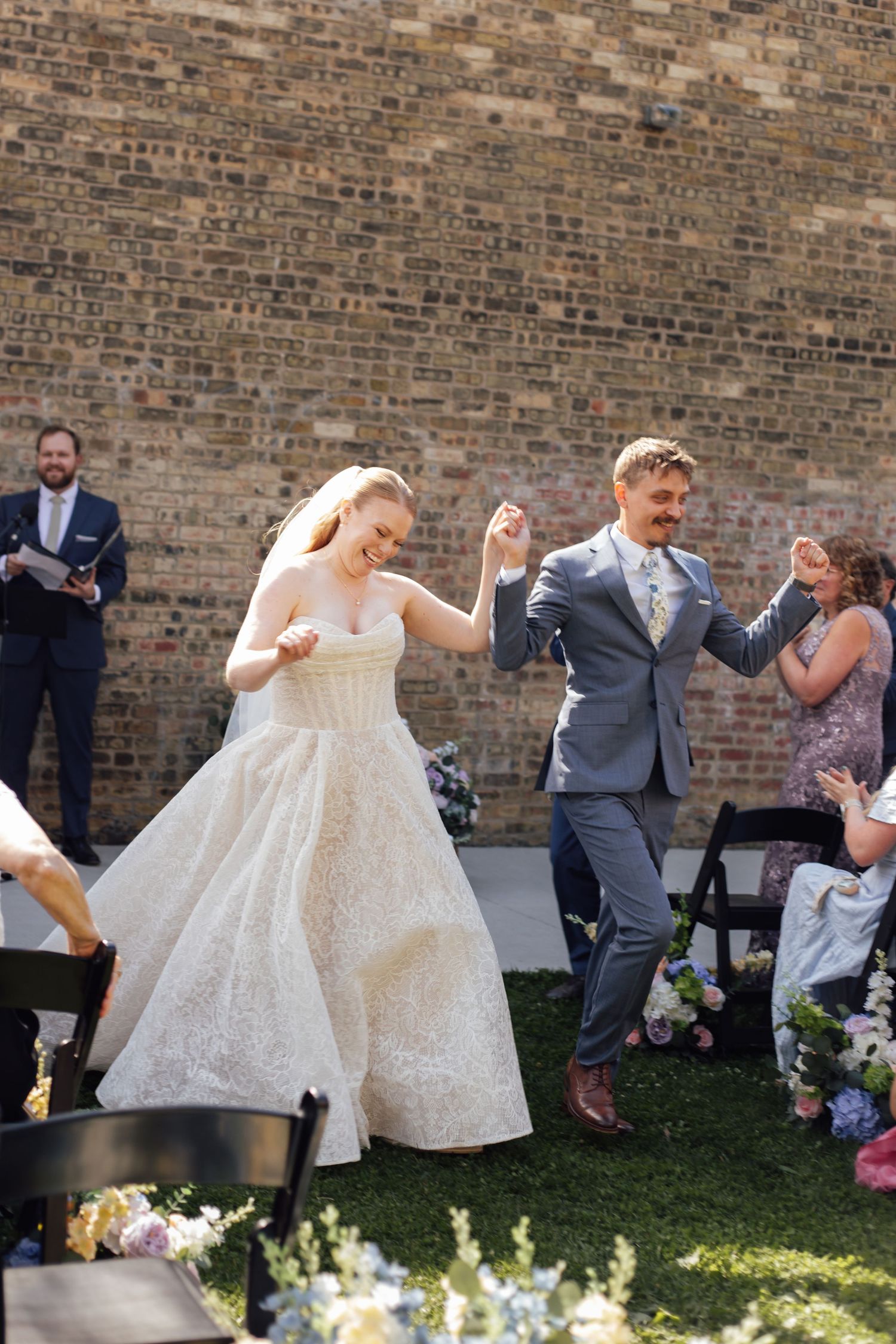 Joyful newlyweds walk back down the aisle after their outdoor wedding ceremony in front of a brick wall.