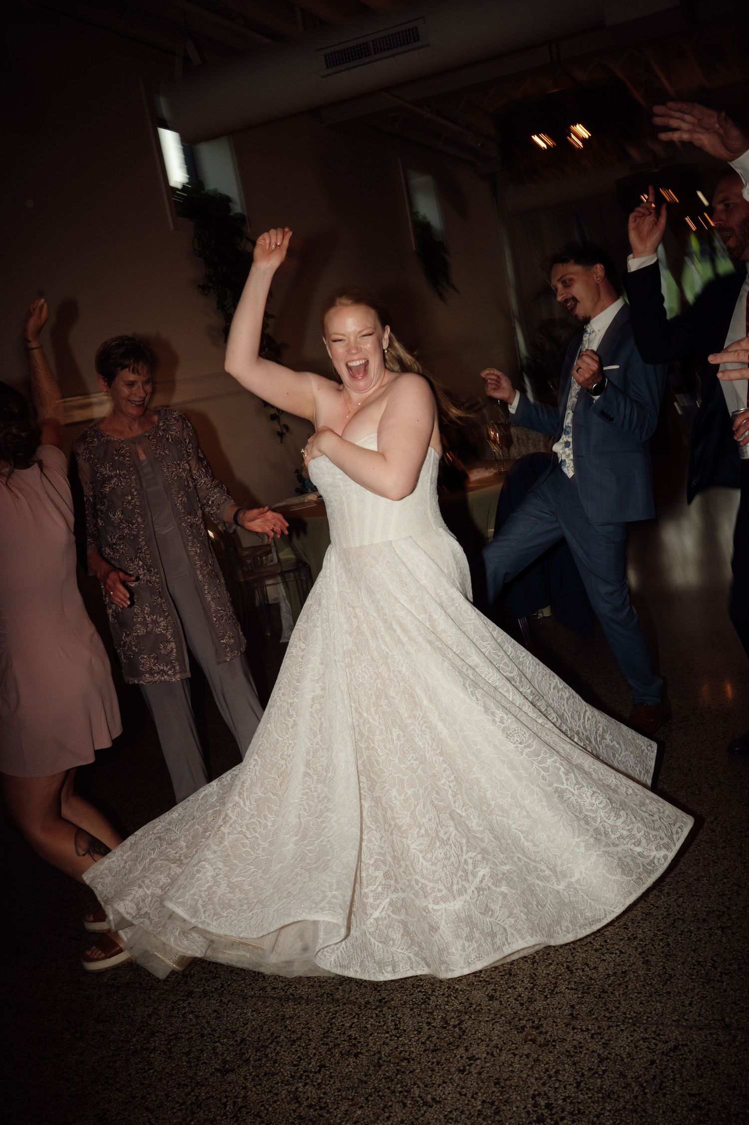 Bride in white lace gown dancing joyfully with arms raised at wedding reception surrounded by guests.