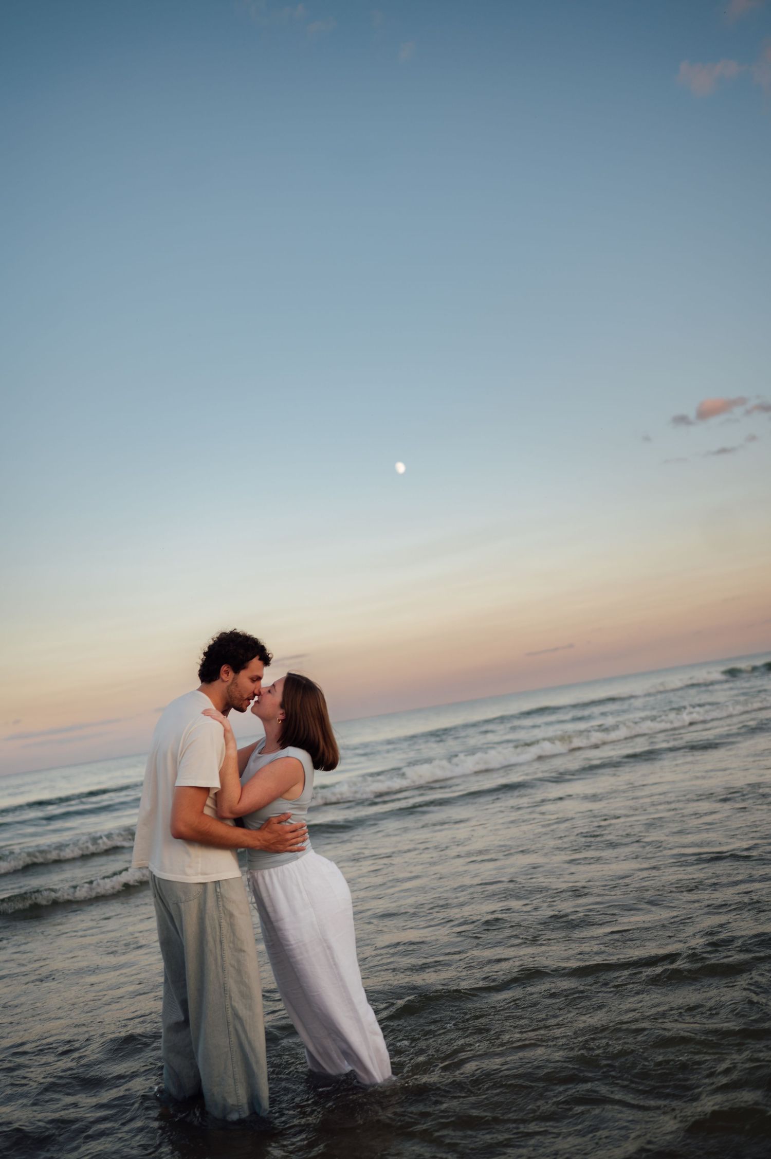 Couple kissing at sunset in ocean waves, wearing white outfits with moon visible in pastel sky.