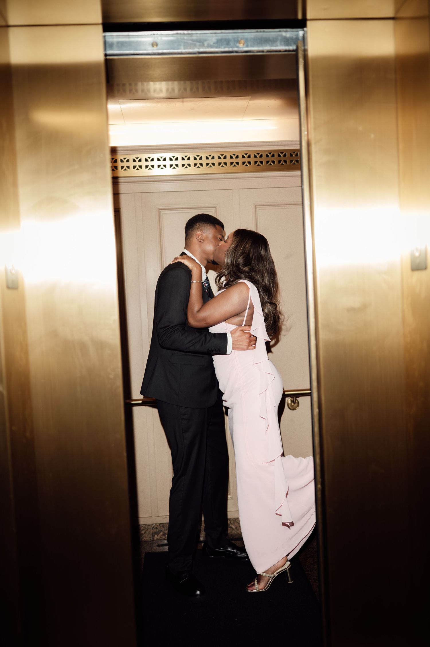 Couple sharing a romantic kiss in a golden elevator, man in black suit and woman in elegant white dress.