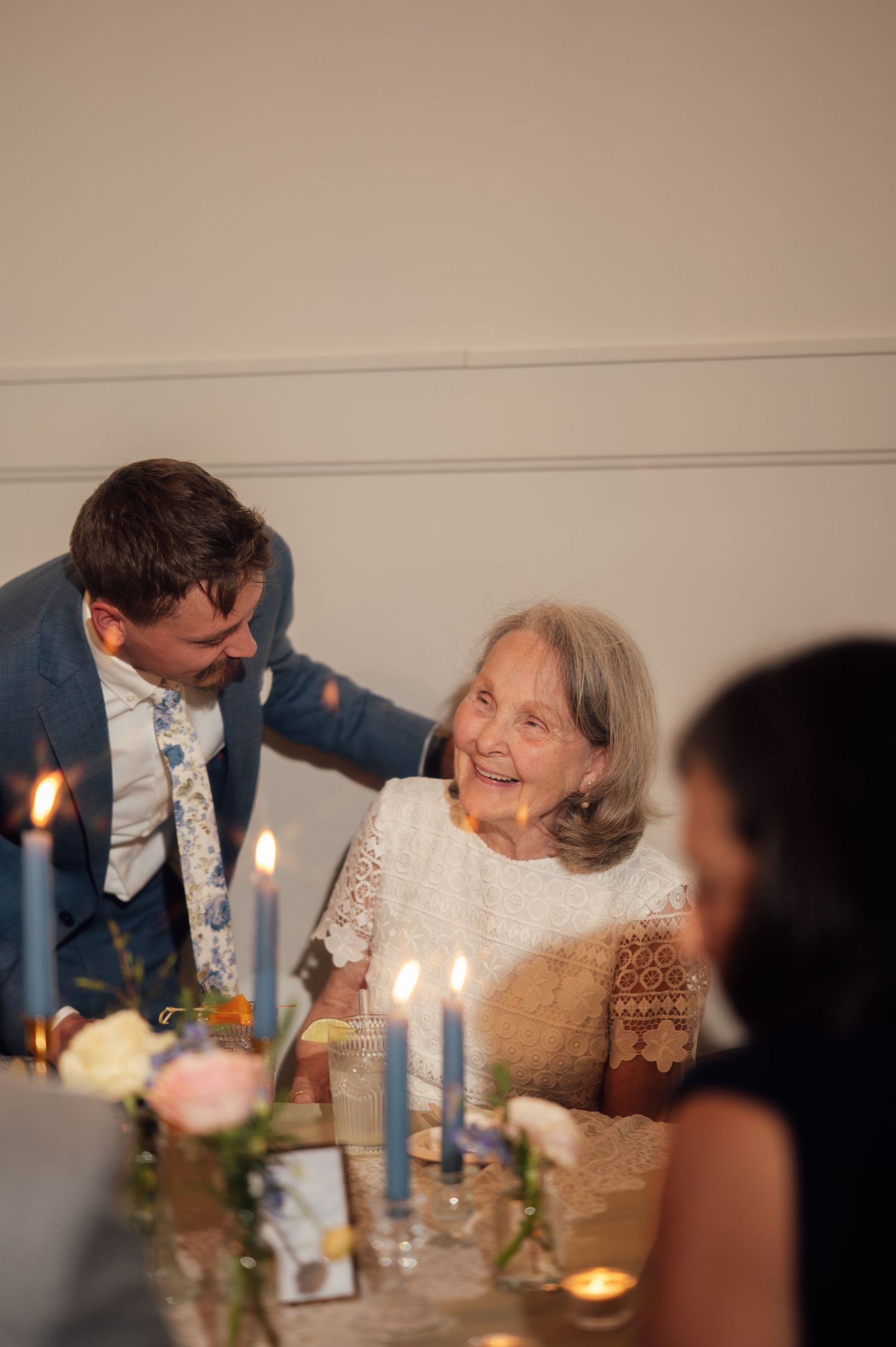 Elderly woman smiling warmly at a candlelit wedding reception table with a man in a blue suit beside her.