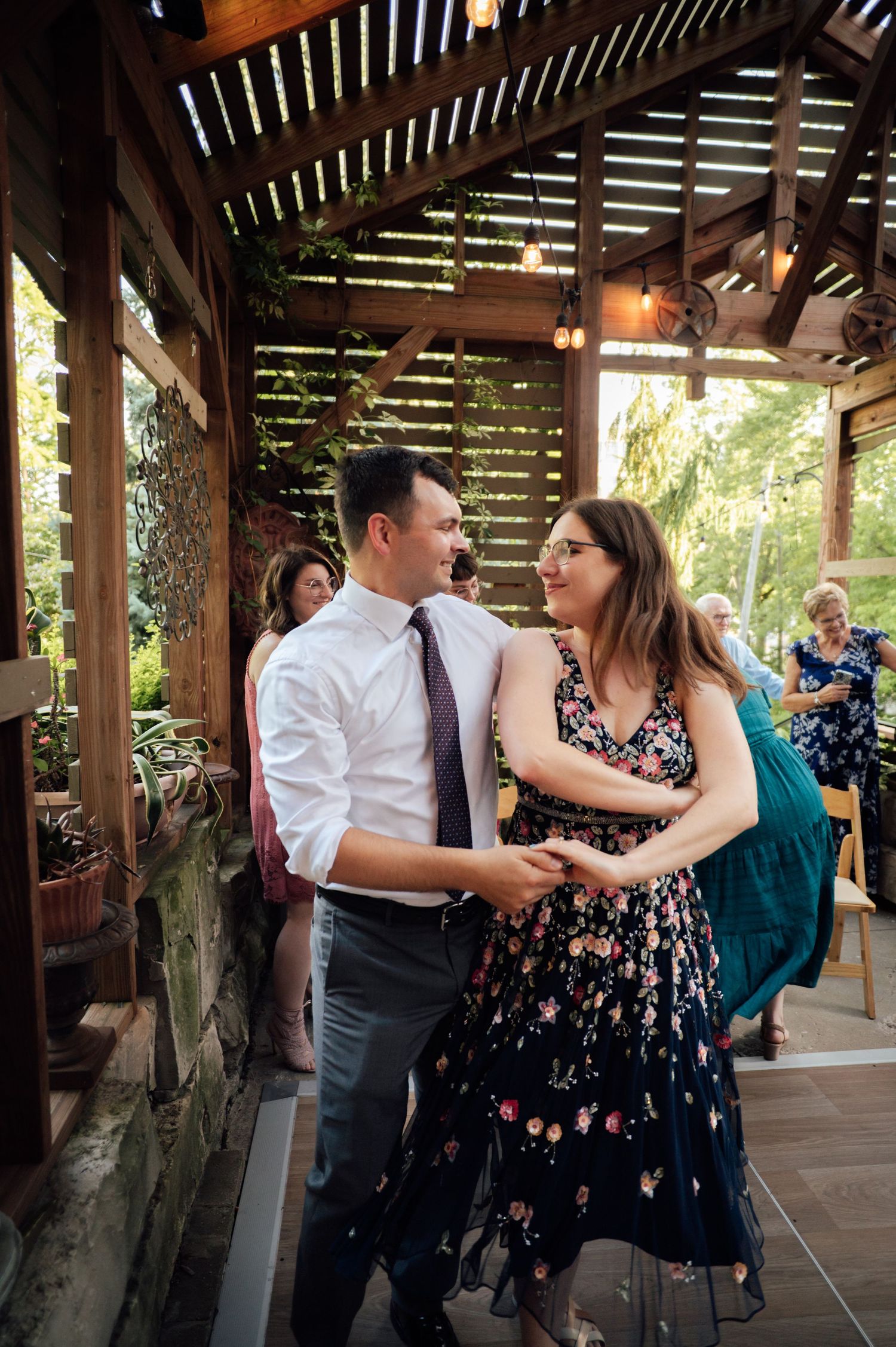 Couple dancing and smiling at an outdoor wedding reception under a wooden pergola with string lights.