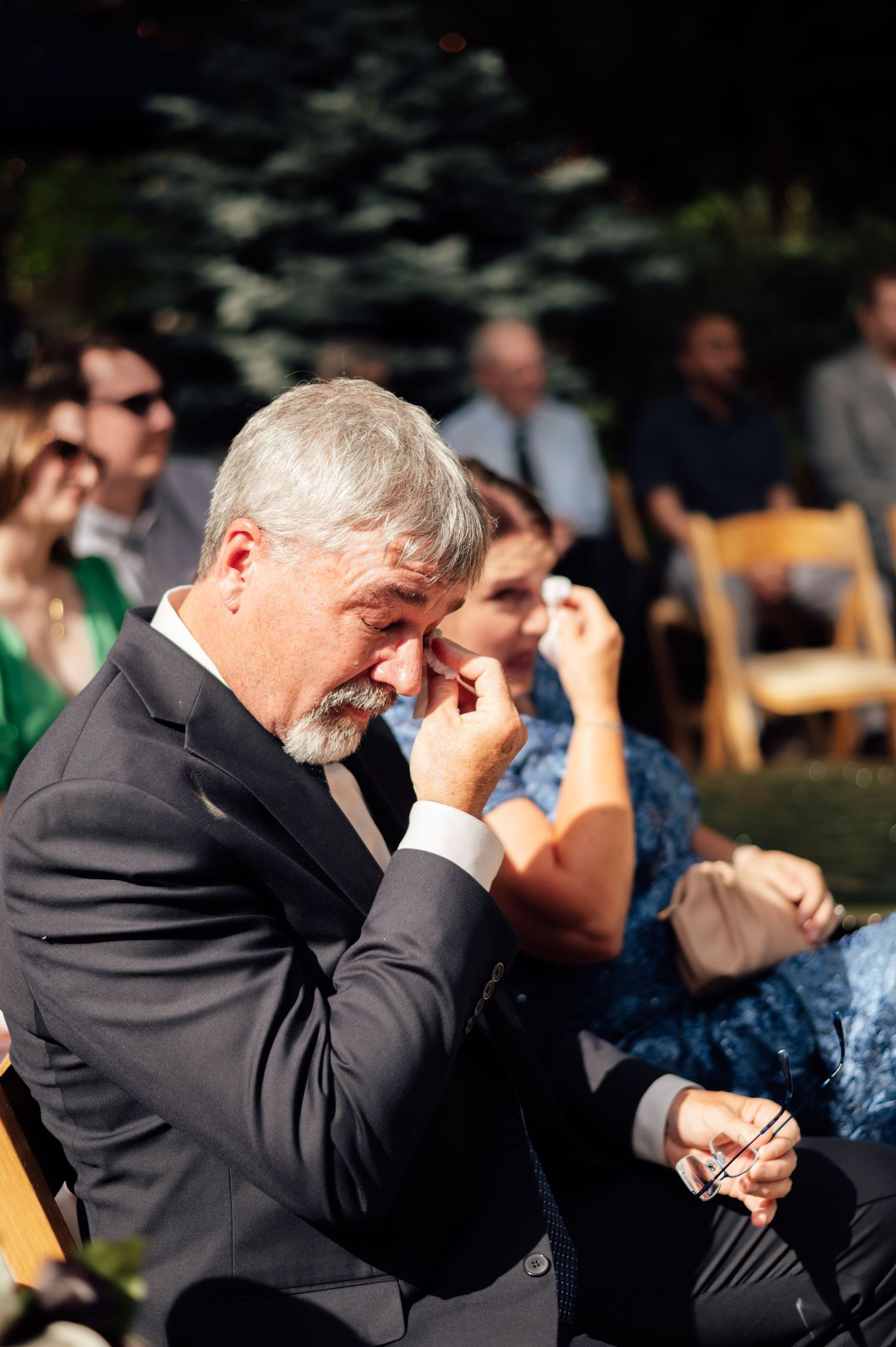 Emotional wedding guest wiping tears during outdoor ceremony, seated among other guests.