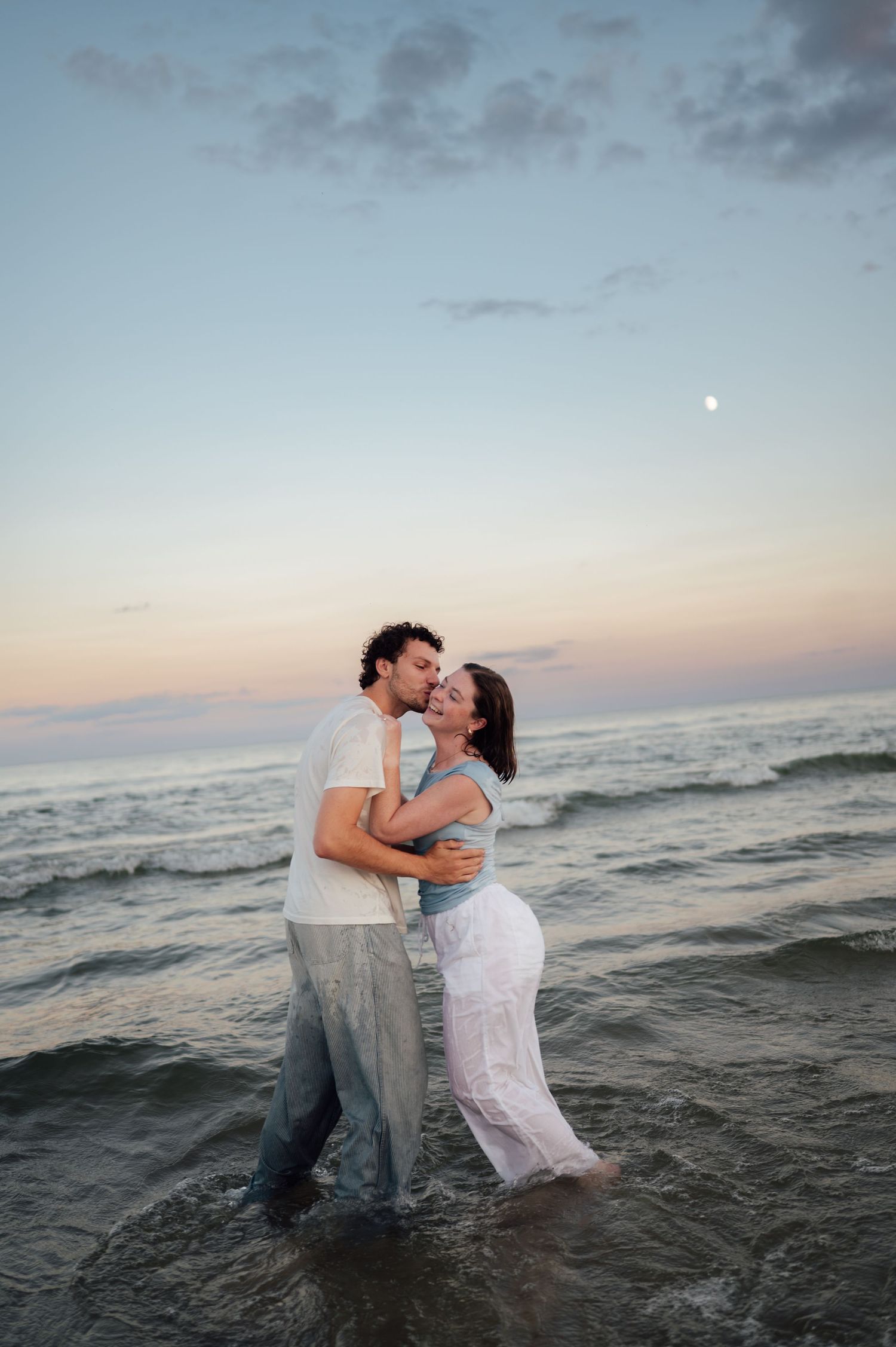 Romantic couple embracing in ocean waves at dusk, moon glowing above colorful sunset sky.