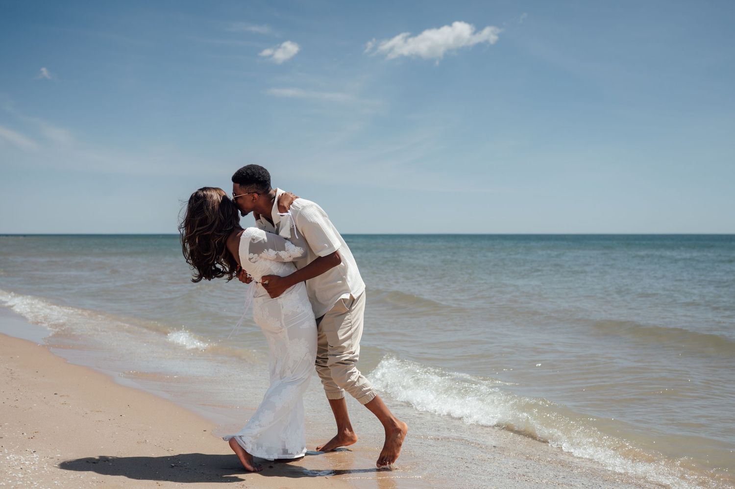 Couple sharing a romantic kiss on a sunny beach, with turquoise ocean waves and blue sky backdrop.