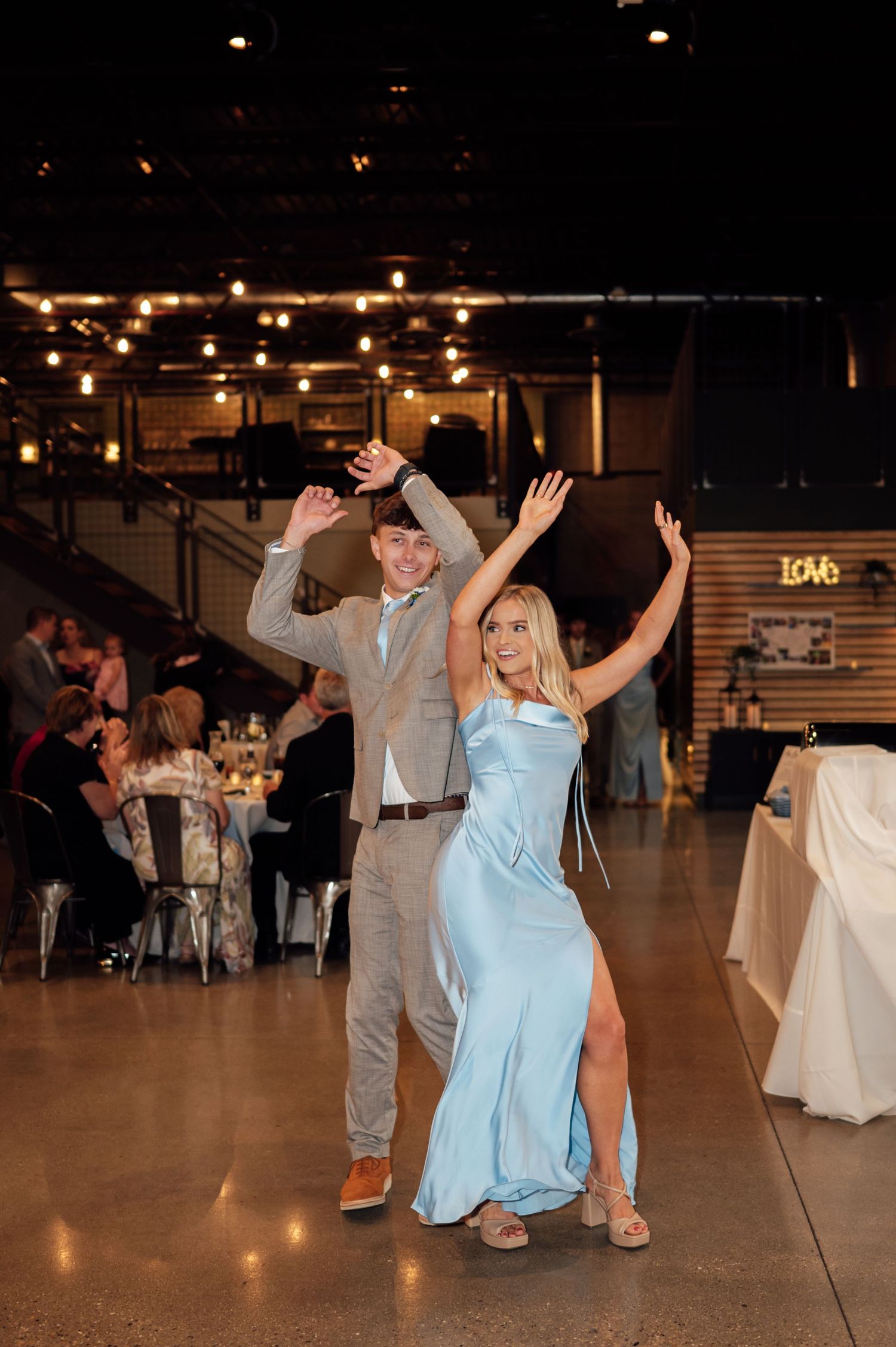 Couple dancing joyfully at wedding reception in elegant venue with string lights, man in gray suit, woman in light blue satin gown.