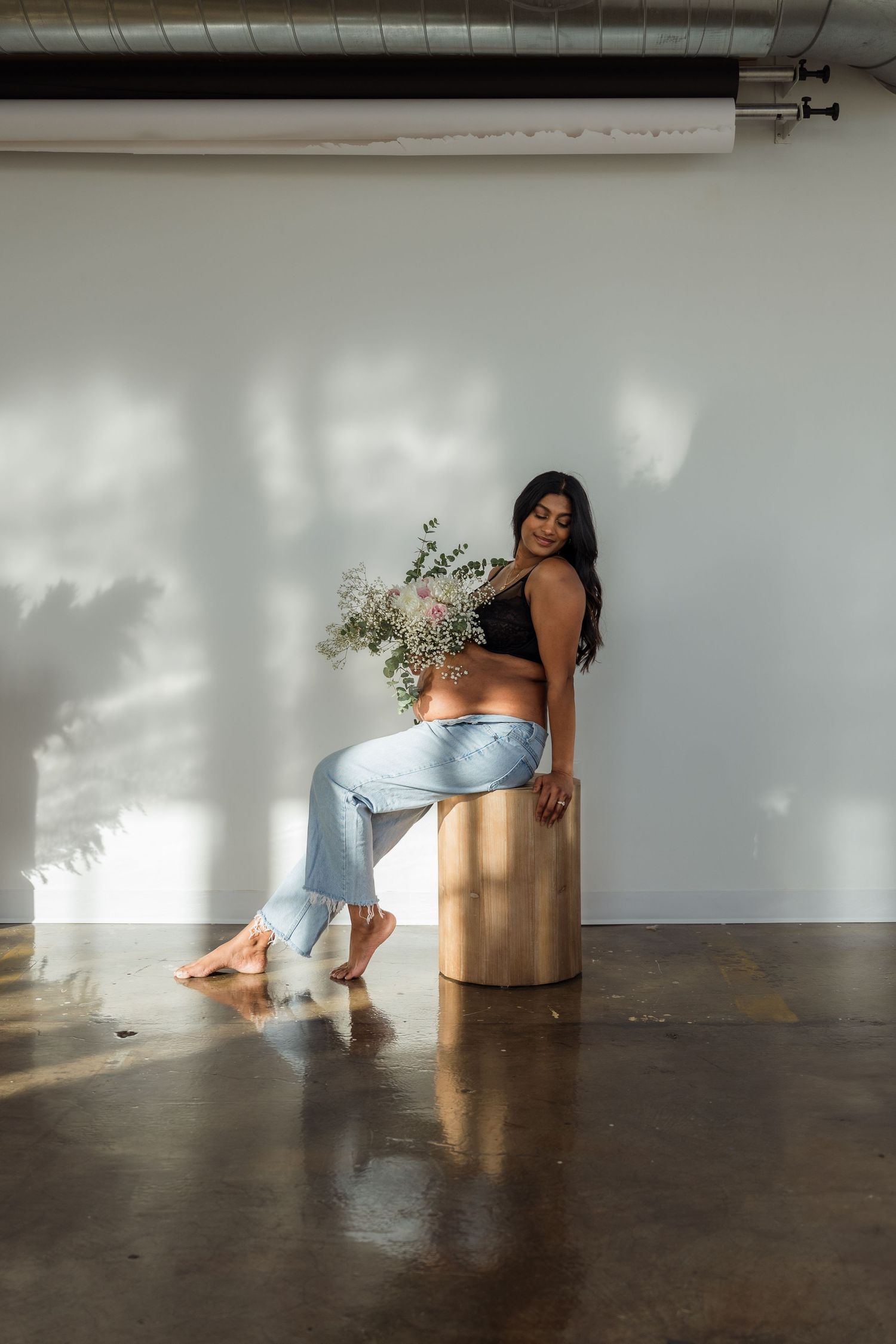 Pregnant woman in black crop top and jeans sits on gold cylinder with flowers.