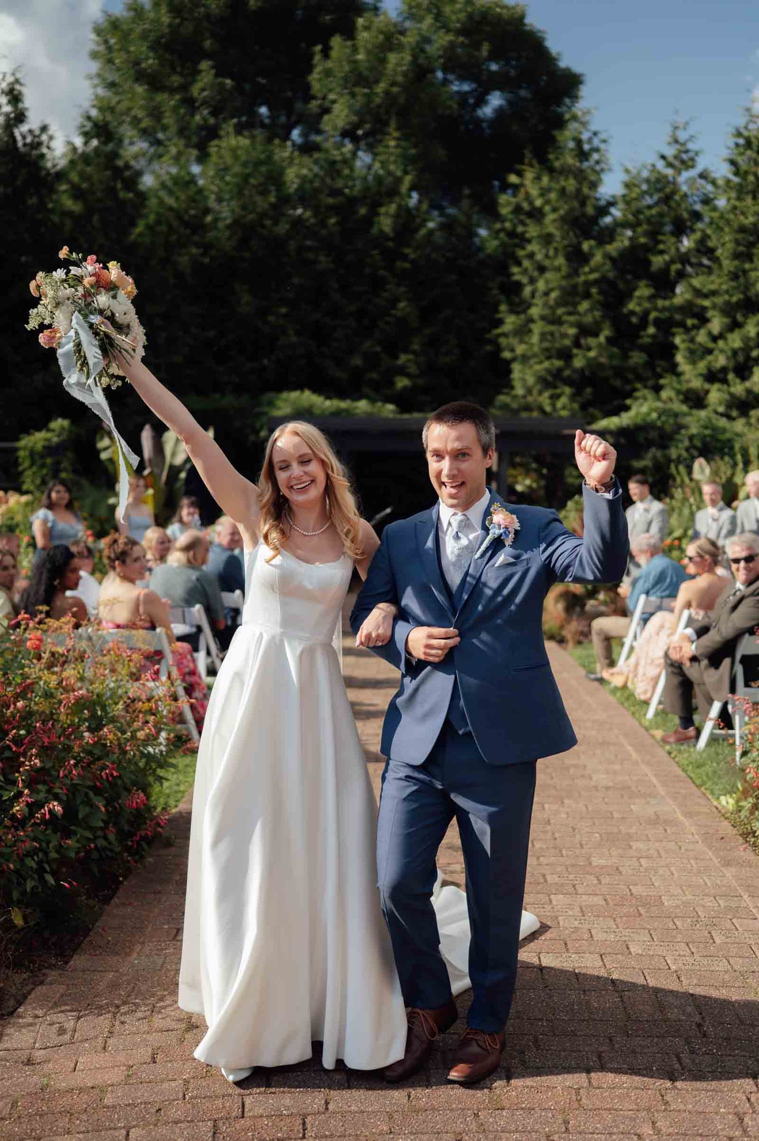 Elated newlyweds raise arms in celebration as they walk back down the aisle after garden wedding.