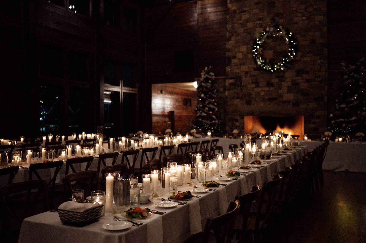 Elegant candlelit wedding reception hall with long tables, Christmas wreaths, and stone fireplace.