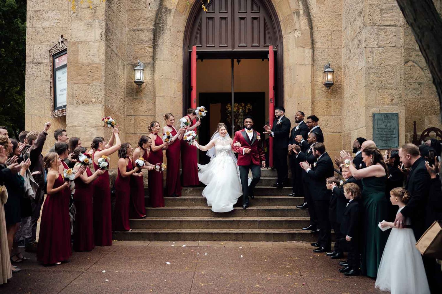 Bride and groom exit a stone church as wedding party lines the steps cheering in burgundy attire.