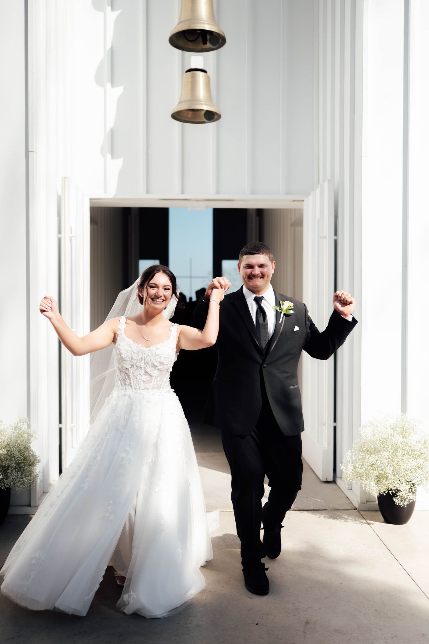 Joyful newlyweds celebrate exit from white chapel with raised arms under decorative bell at ceremony entrance.