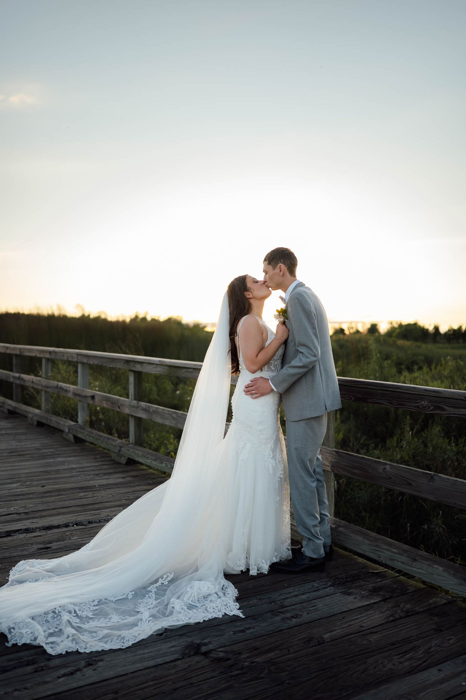 Bride and groom share a romantic sunset kiss on a wooden boardwalk surrounded by greenery.
