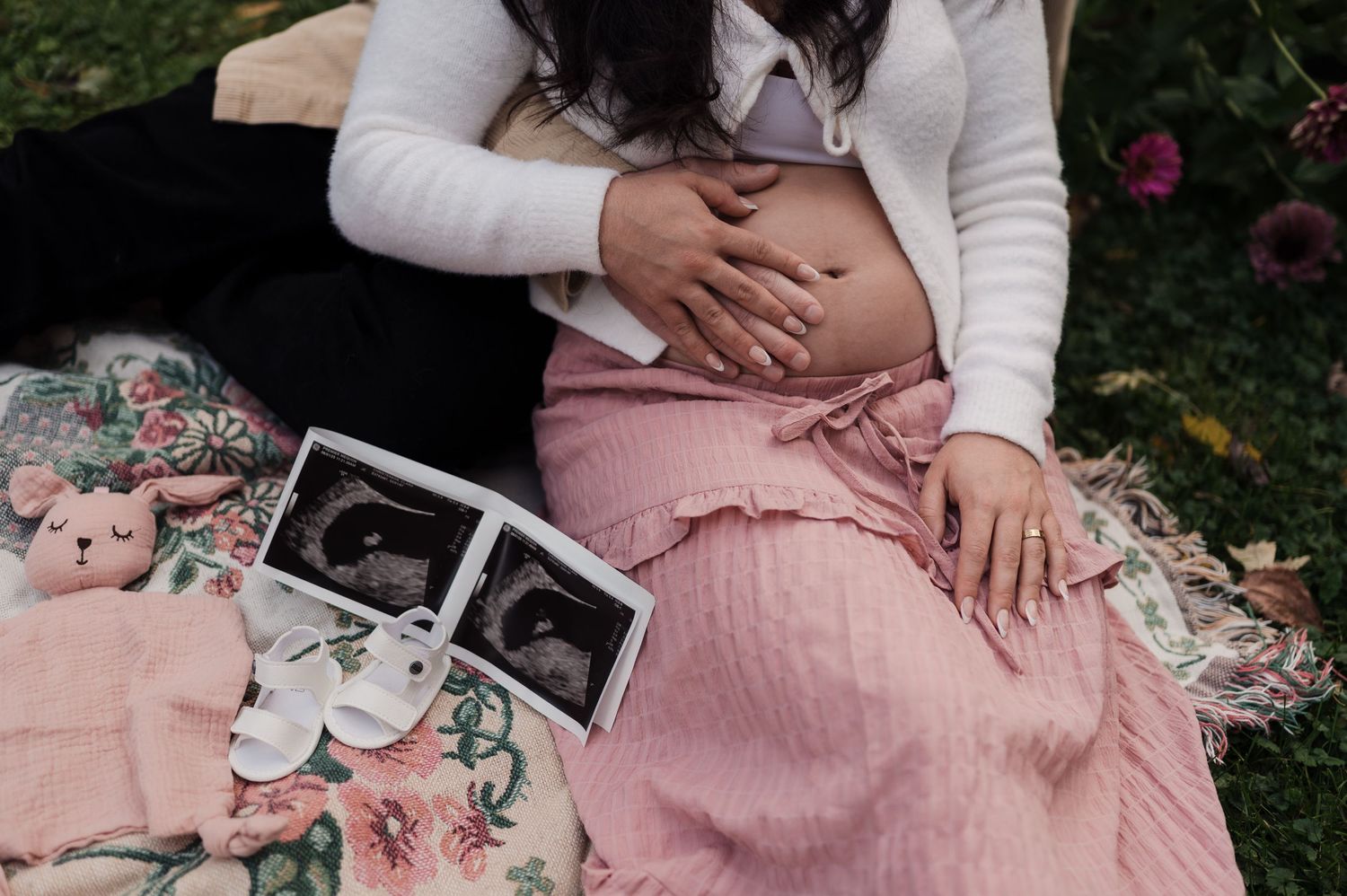 Pregnant woman sitting on floral blanket with ultrasound photos and baby shoes beside her.