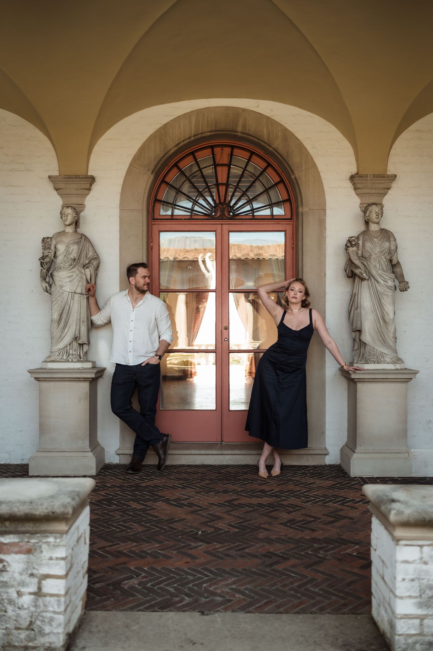 Elegant couple posing between classical statues under arched portico with ornate red double doors.