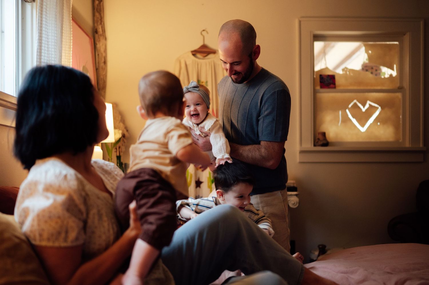 A bald father smiles at his laughing baby while the mother watches lovingly in a cozy bedroom.