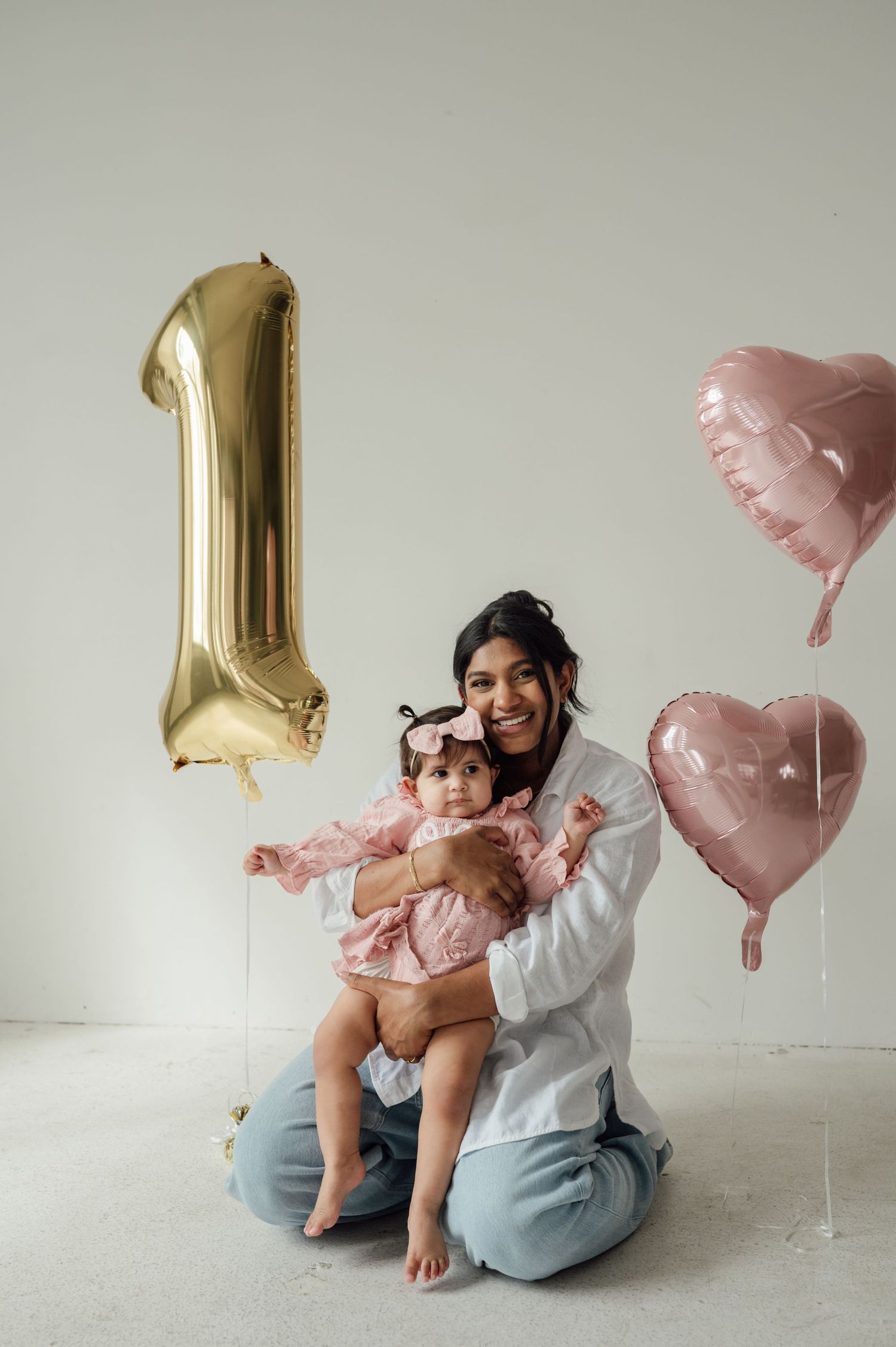 Mother sitting on floor holding baby girl in pink dress with gold number one birthday balloons.