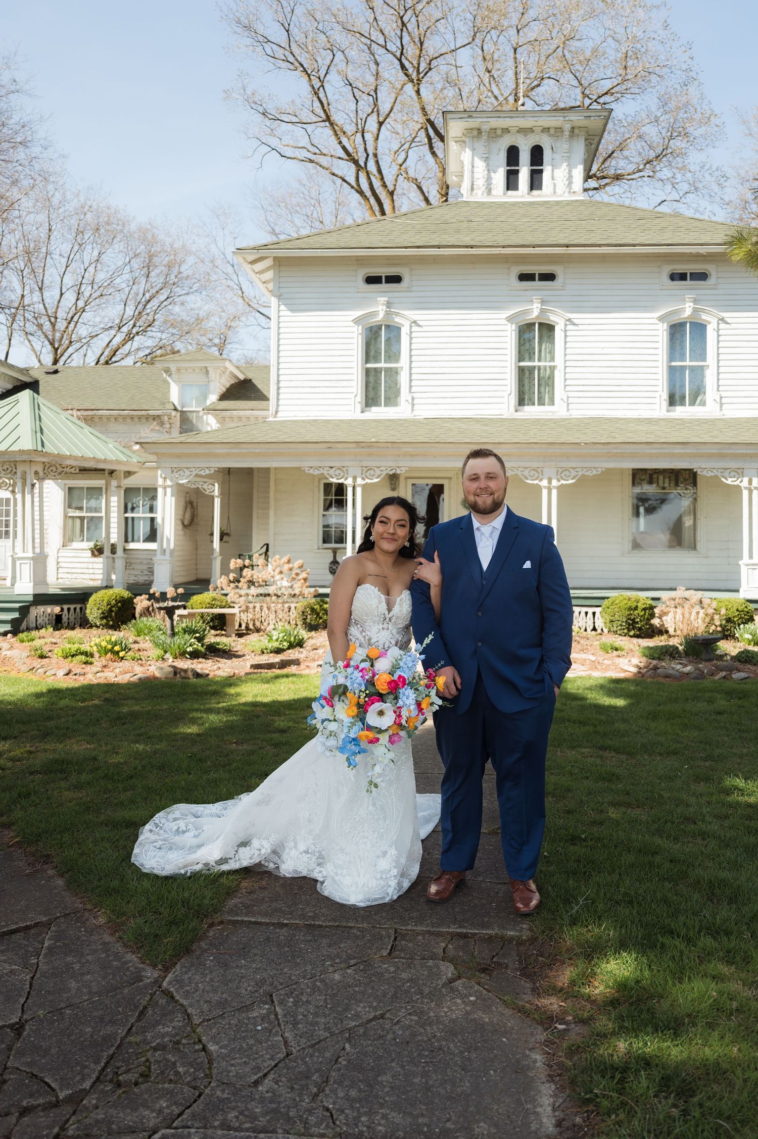 A couple stands in wedding attire in front of a beautiful white Victorian-style house with a wraparound porch.