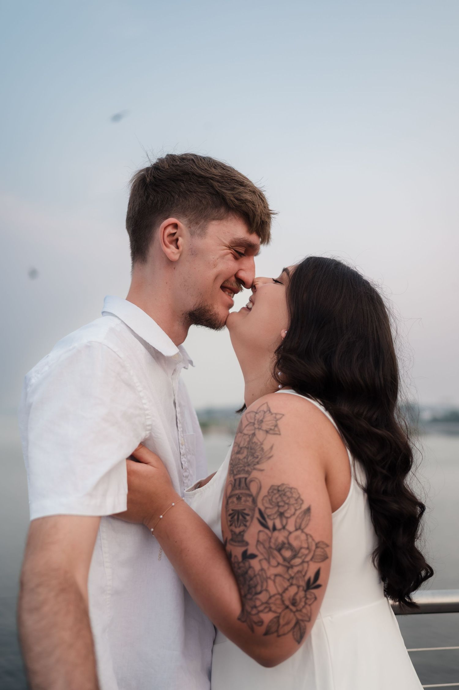 A series of romantic waterfront wedding photos showing a couple in white embracing with visible tattoo artwork.
