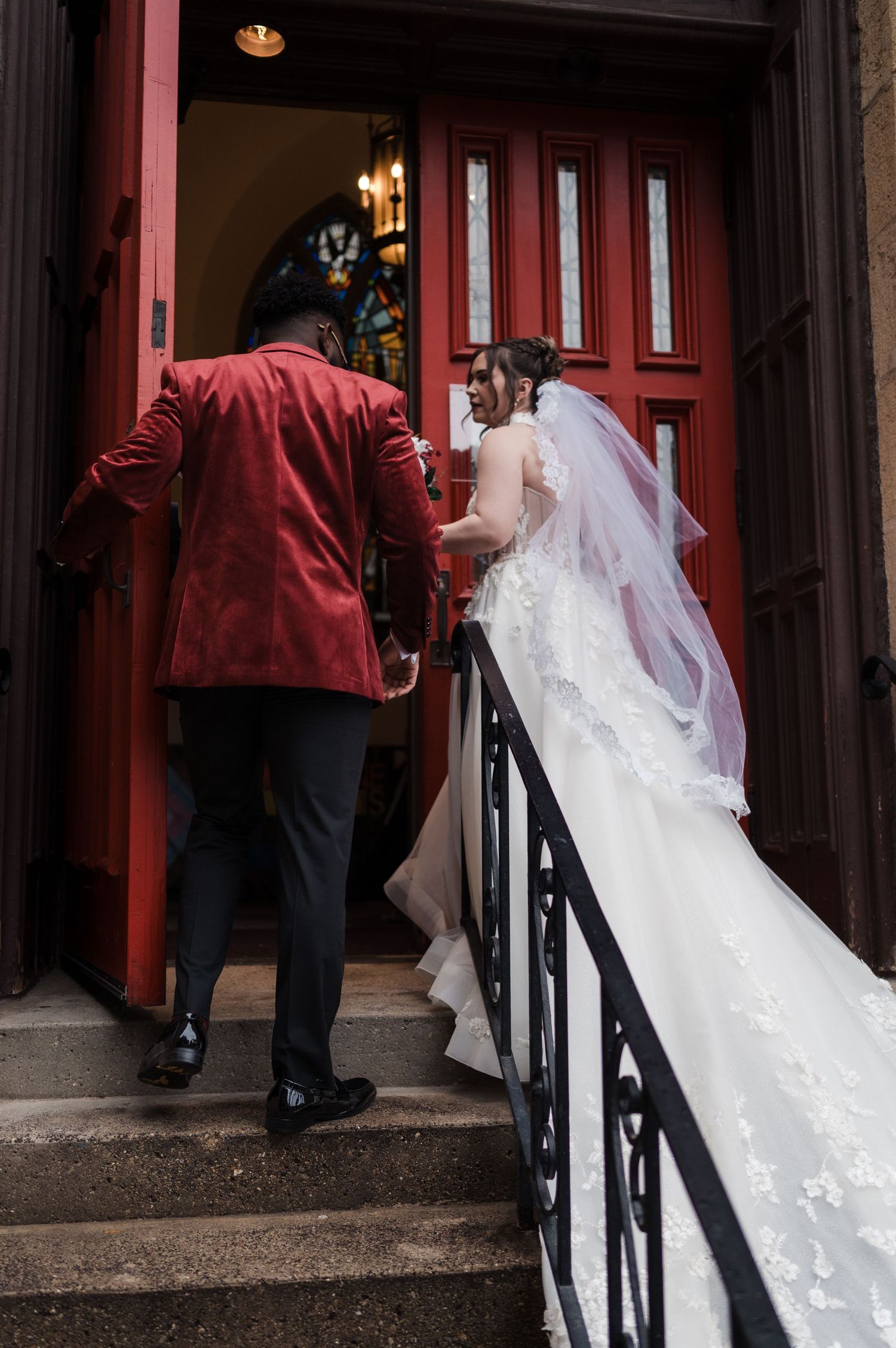 A bride's flowing white veil trails behind as she enters a doorway with red walls and black railings.