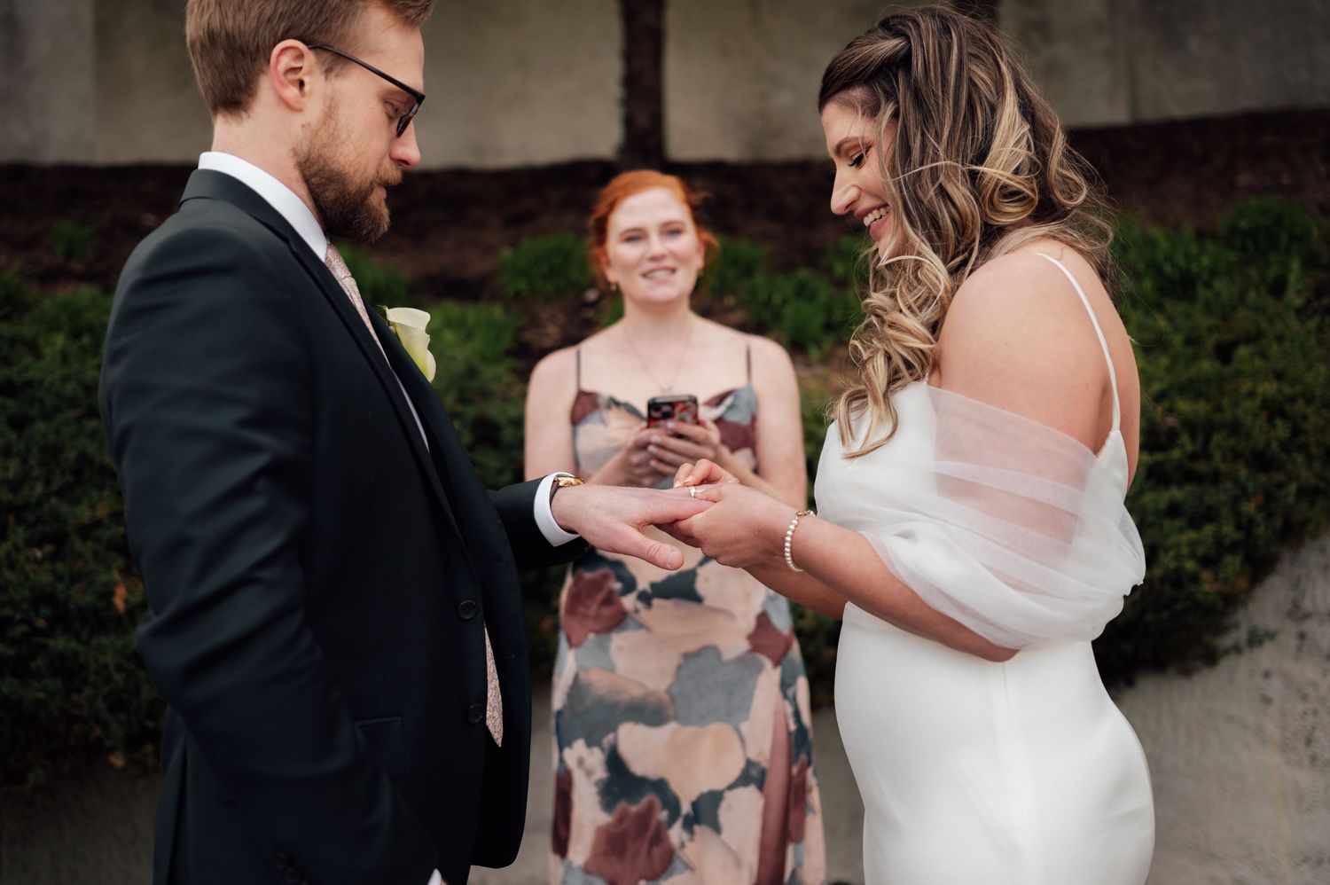Bride and groom exchanging rings during outdoor wedding ceremony with smiling officiant in floral dress in background.