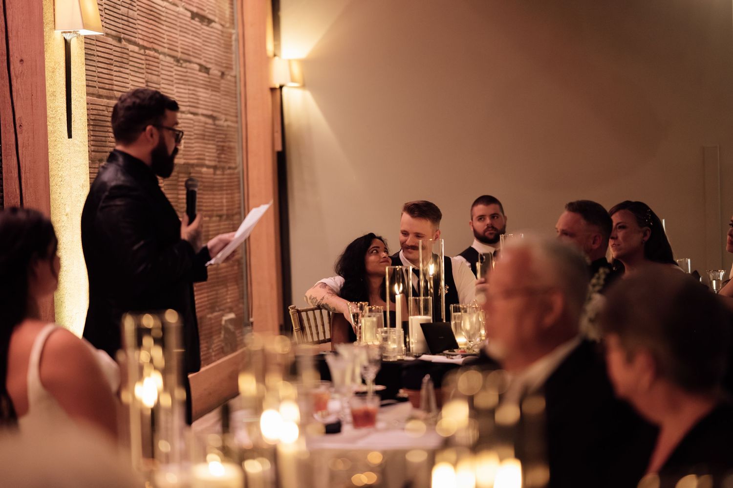 Man giving a speech at an elegant candlelit wedding reception dinner with guests seated at decorated tables.