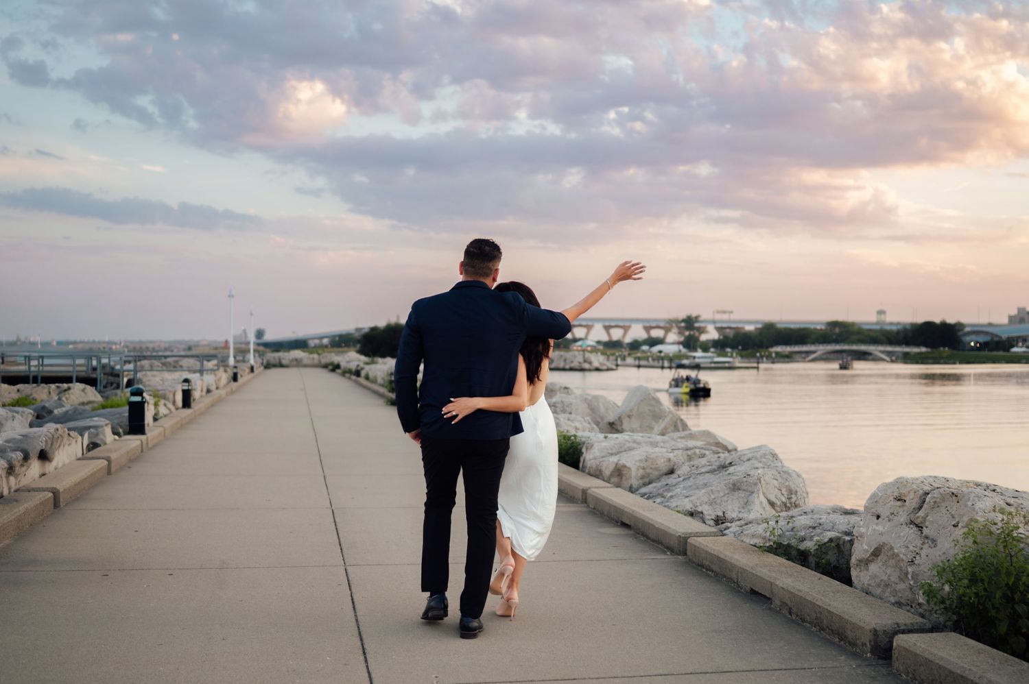 Couple walking along waterfront promenade at sunset, woman pointing toward horizon, man in navy suit, white dress.