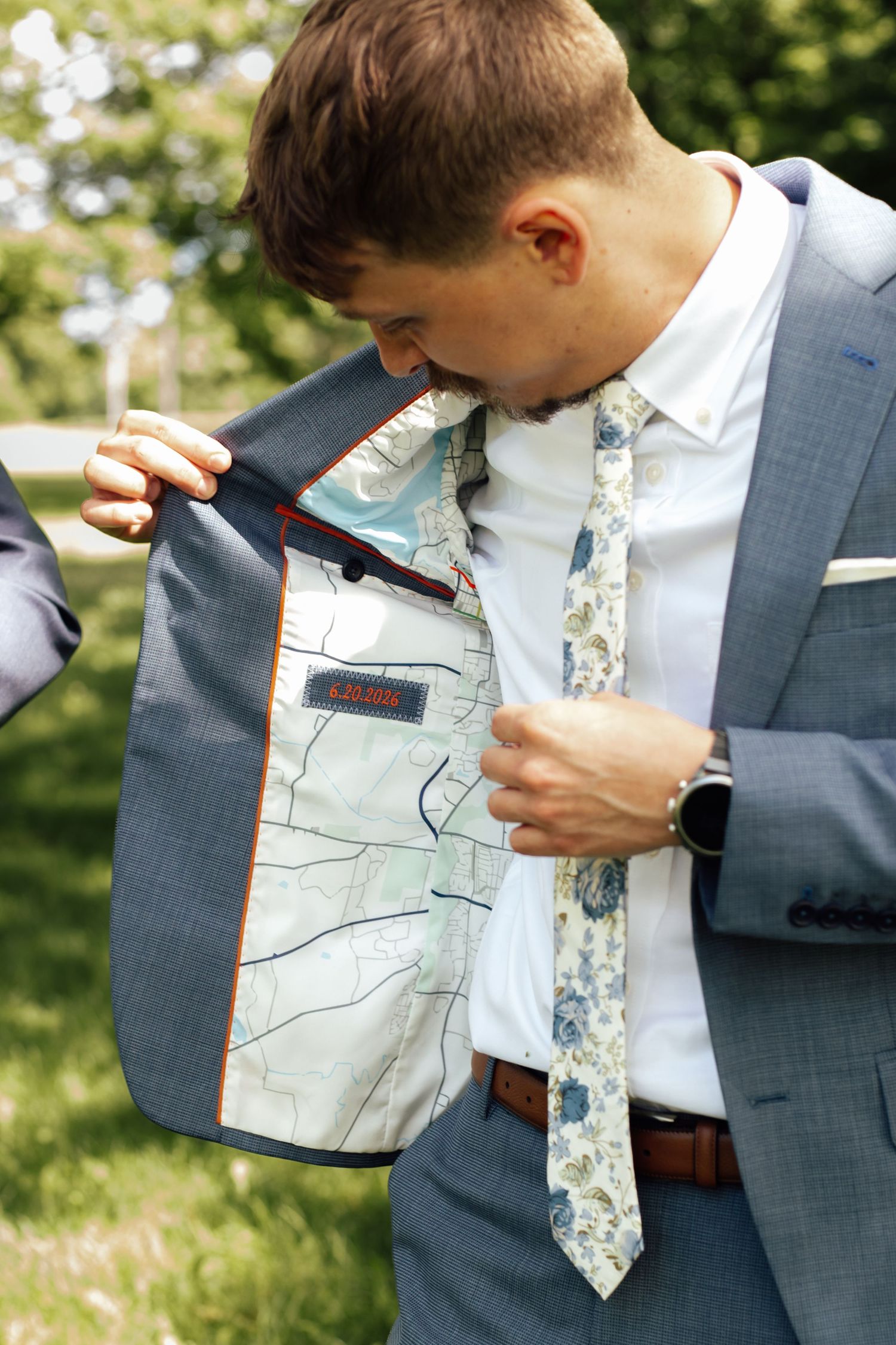 Man in gray suit opens jacket revealing map-print lining, wearing floral tie and white shirt outdoors.