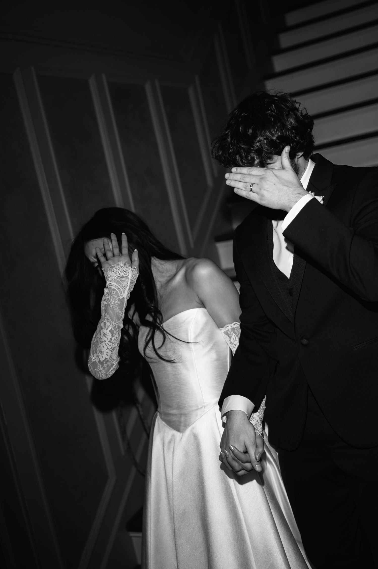 Black and white photo of bride and groom playfully covering their faces while holding hands.