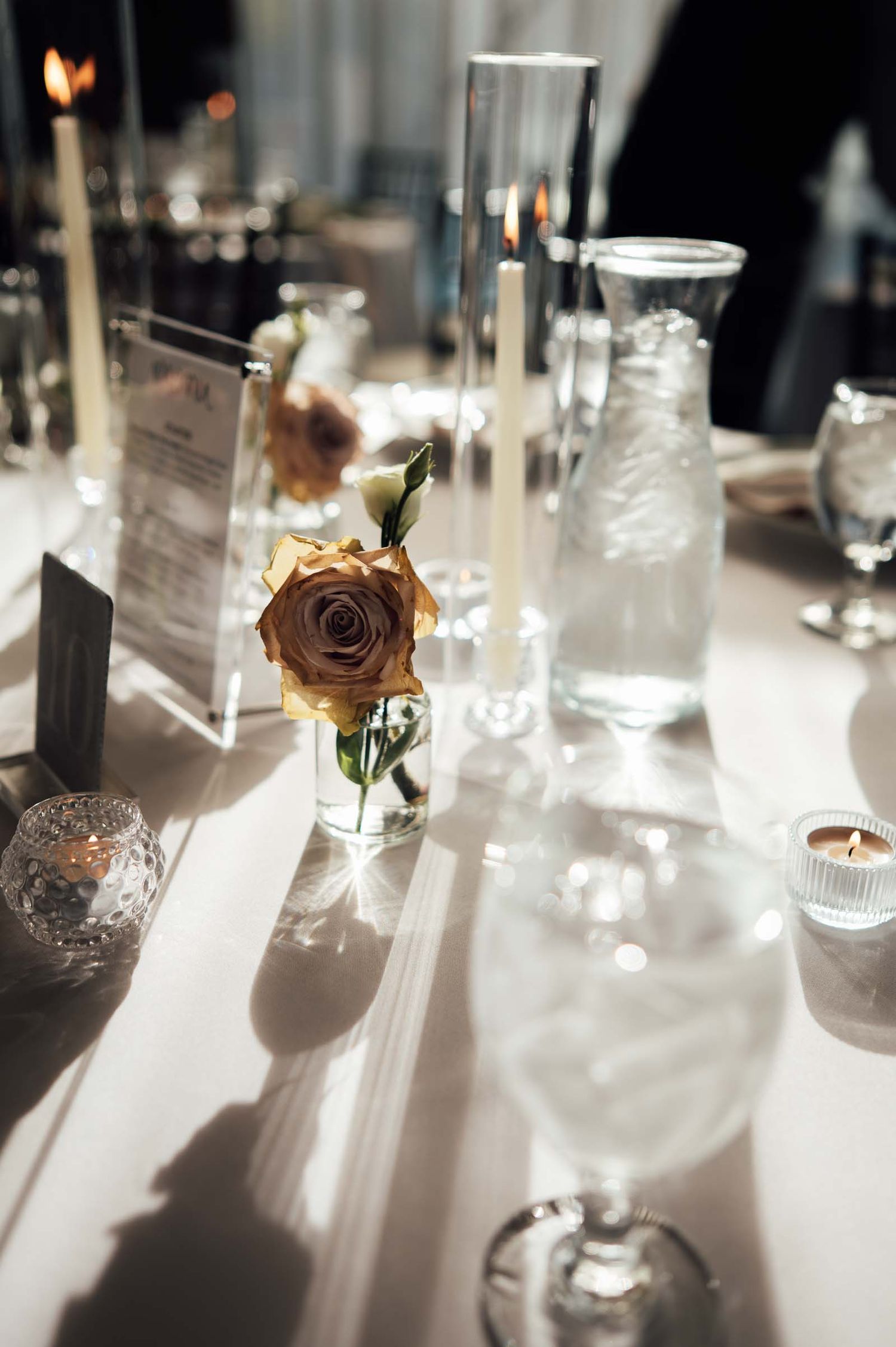 Elegant wedding table centerpiece with a golden rose in a glass vase, candles, and crystal glassware on white linen.