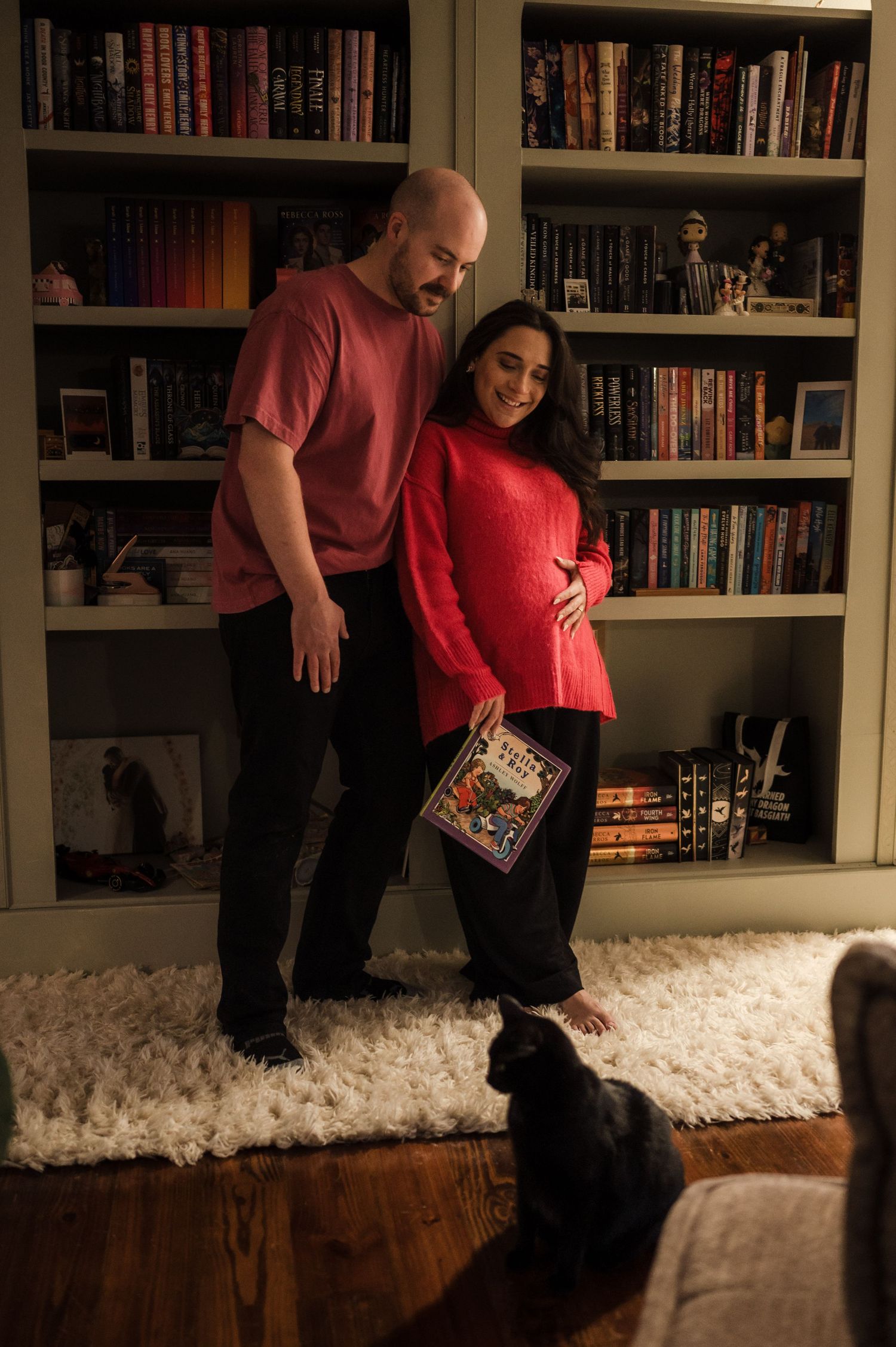 Expecting couple smiling before large bookshelf, woman in red sweater holding book, black cat nearby.