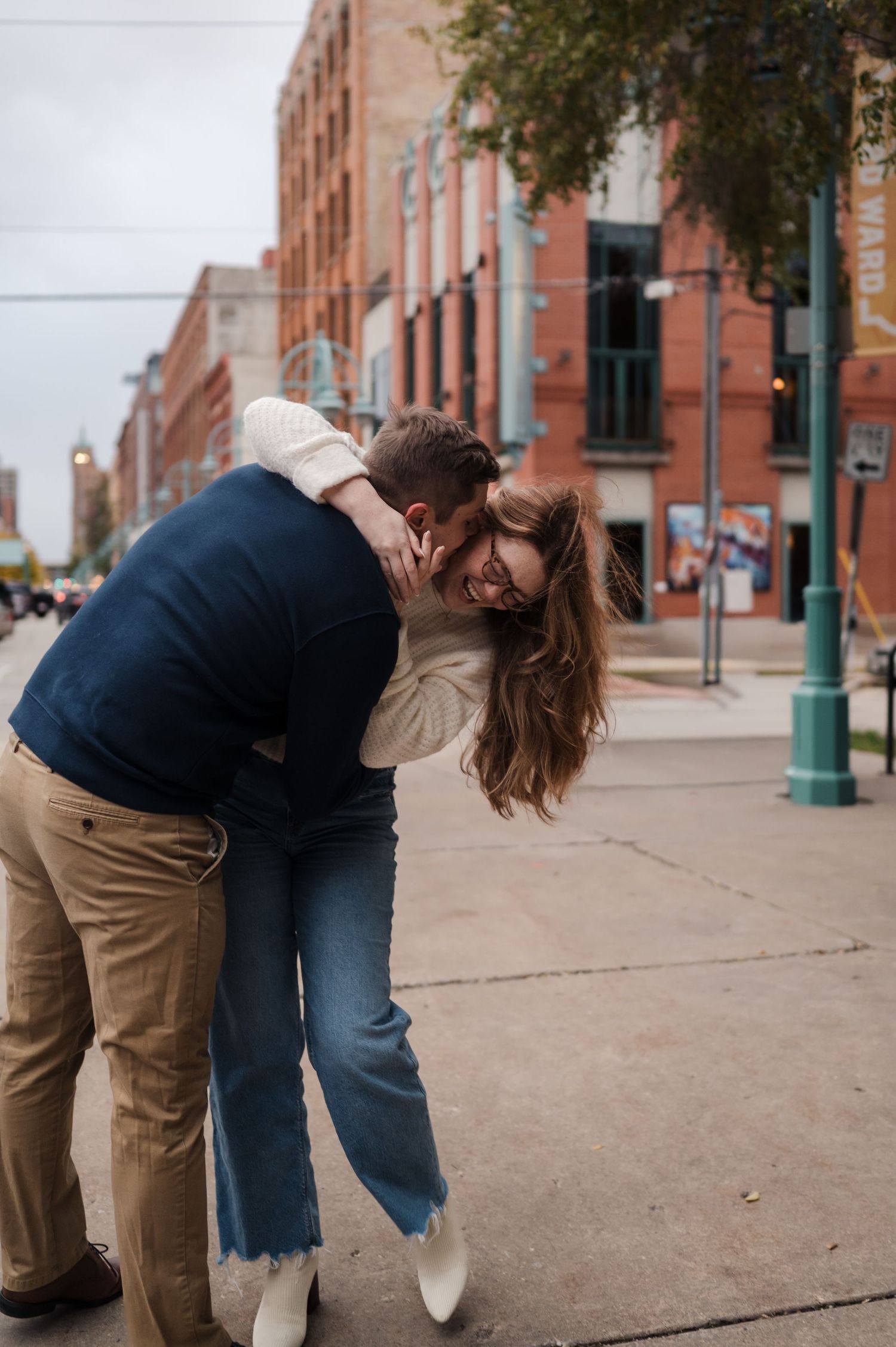 Playful couple laughing and embracing on a city sidewalk with historic brick buildings behind them.