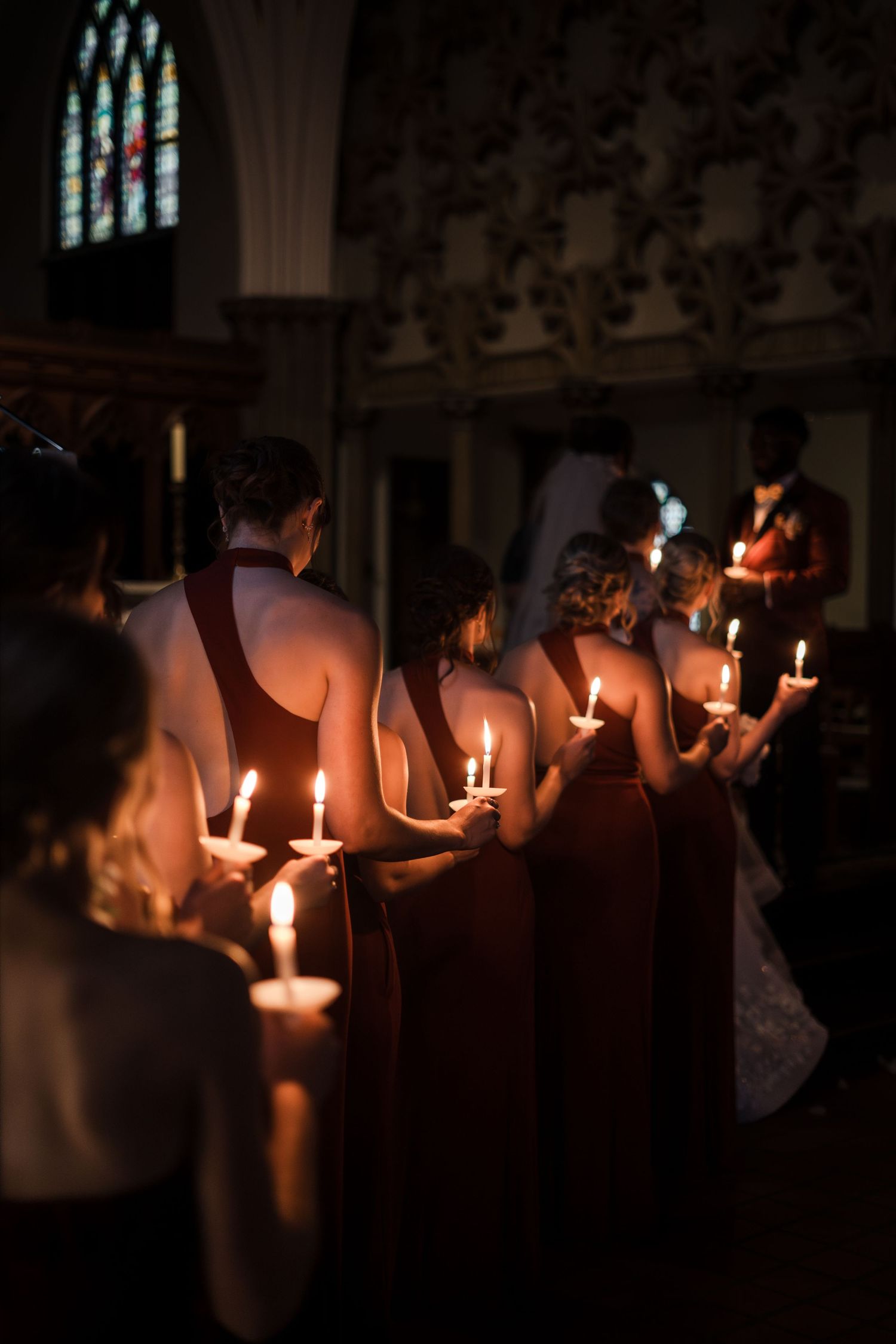 A row of wedding attendees hold glowing candles in a darkened church interior.