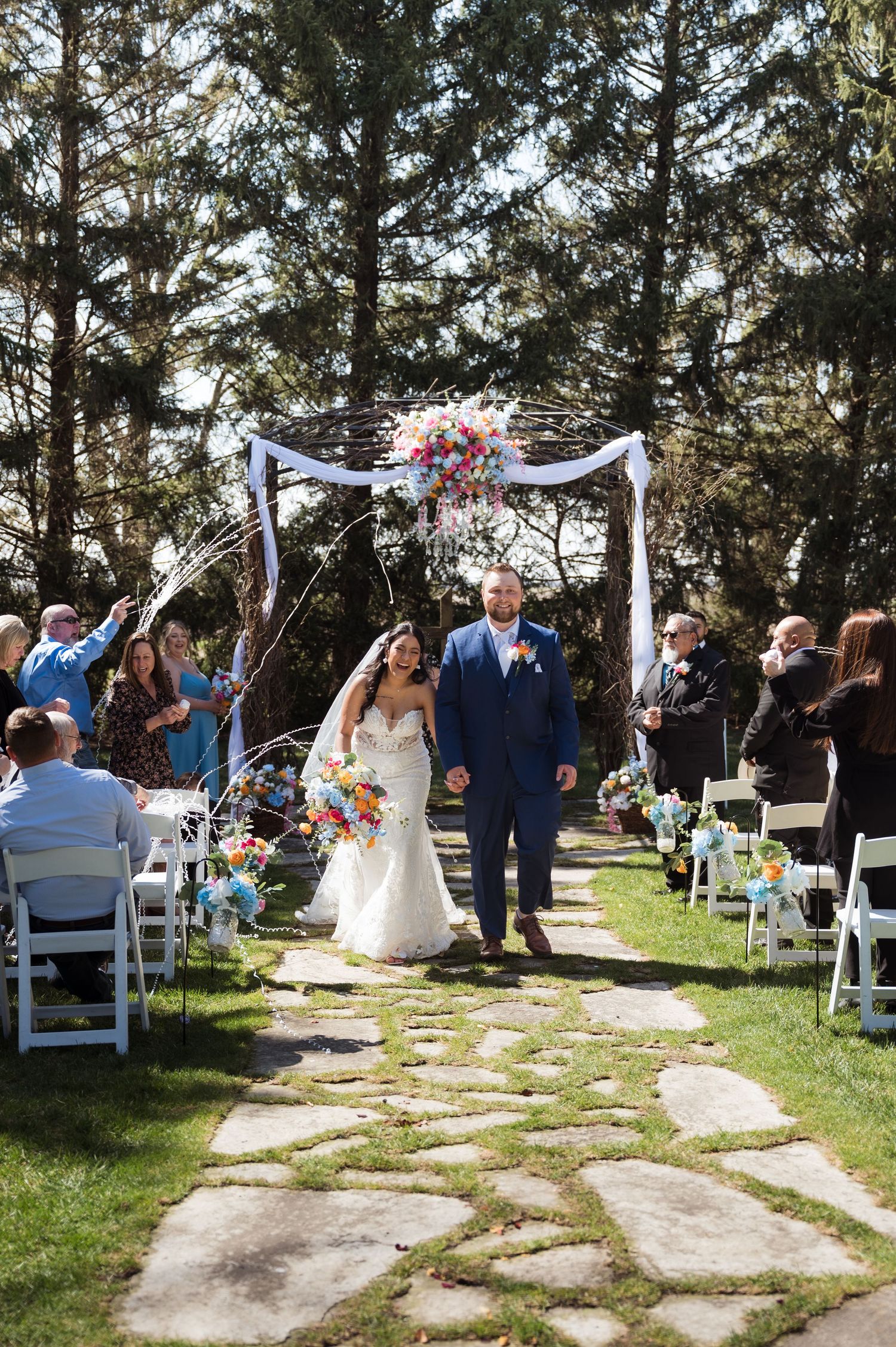 Bride and groom walk down outdoor stone aisle after ceremony under floral arch with colorful blooms.