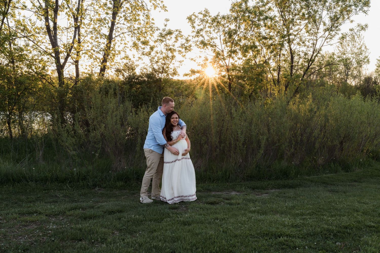 Expecting couple sharing a tender moment at sunset in a green meadow surrounded by trees during a maternity photo session.