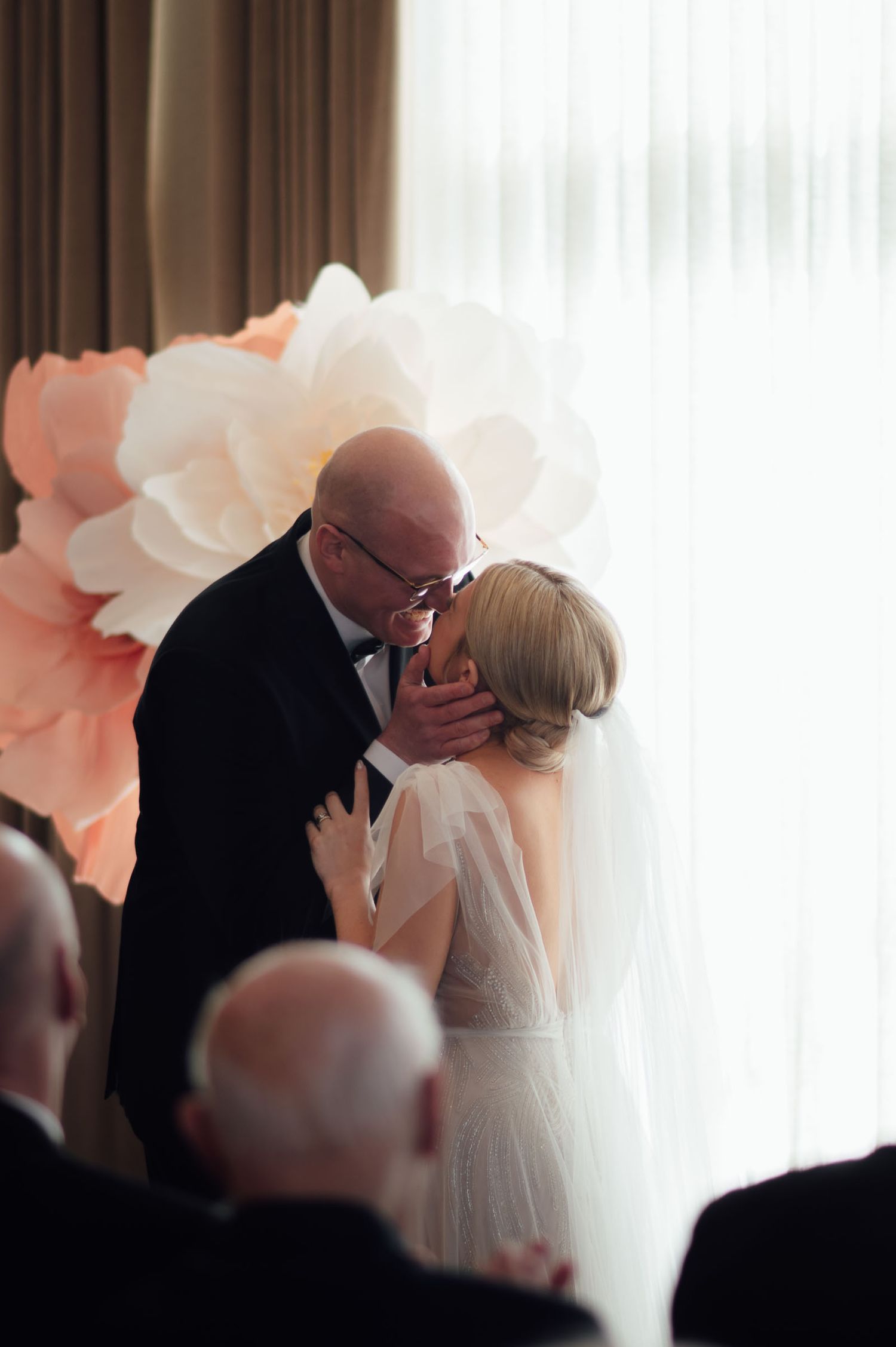 Bride and groom sharing first kiss at altar with large paper flower backdrop at indoor wedding.