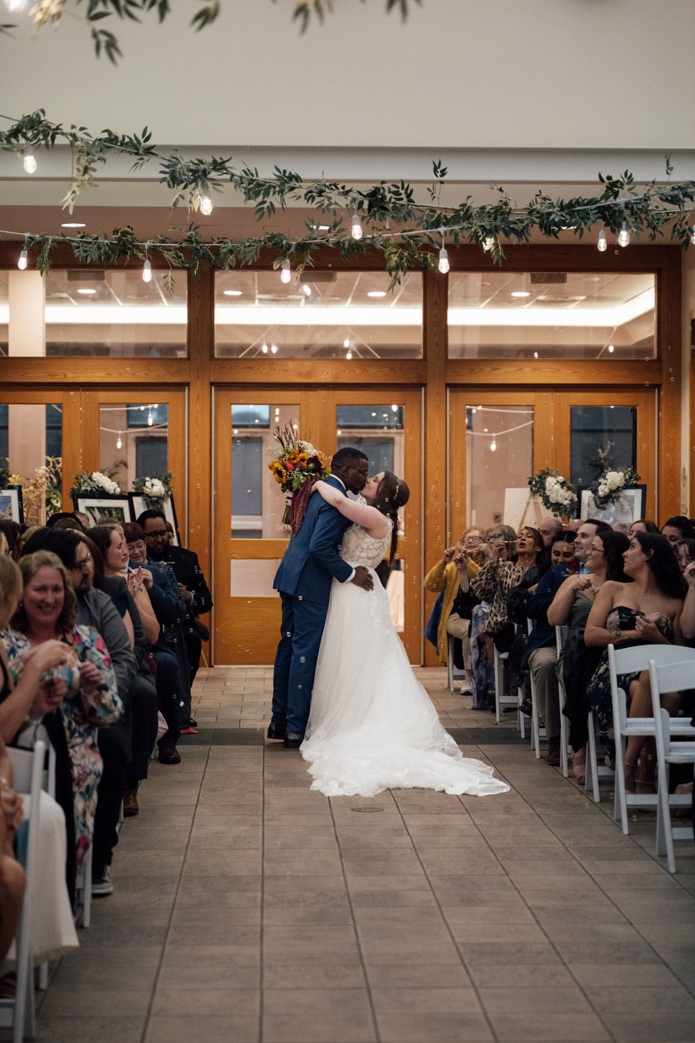 Bride and groom share first kiss at indoor wedding ceremony aisle surrounded by cheering guests.