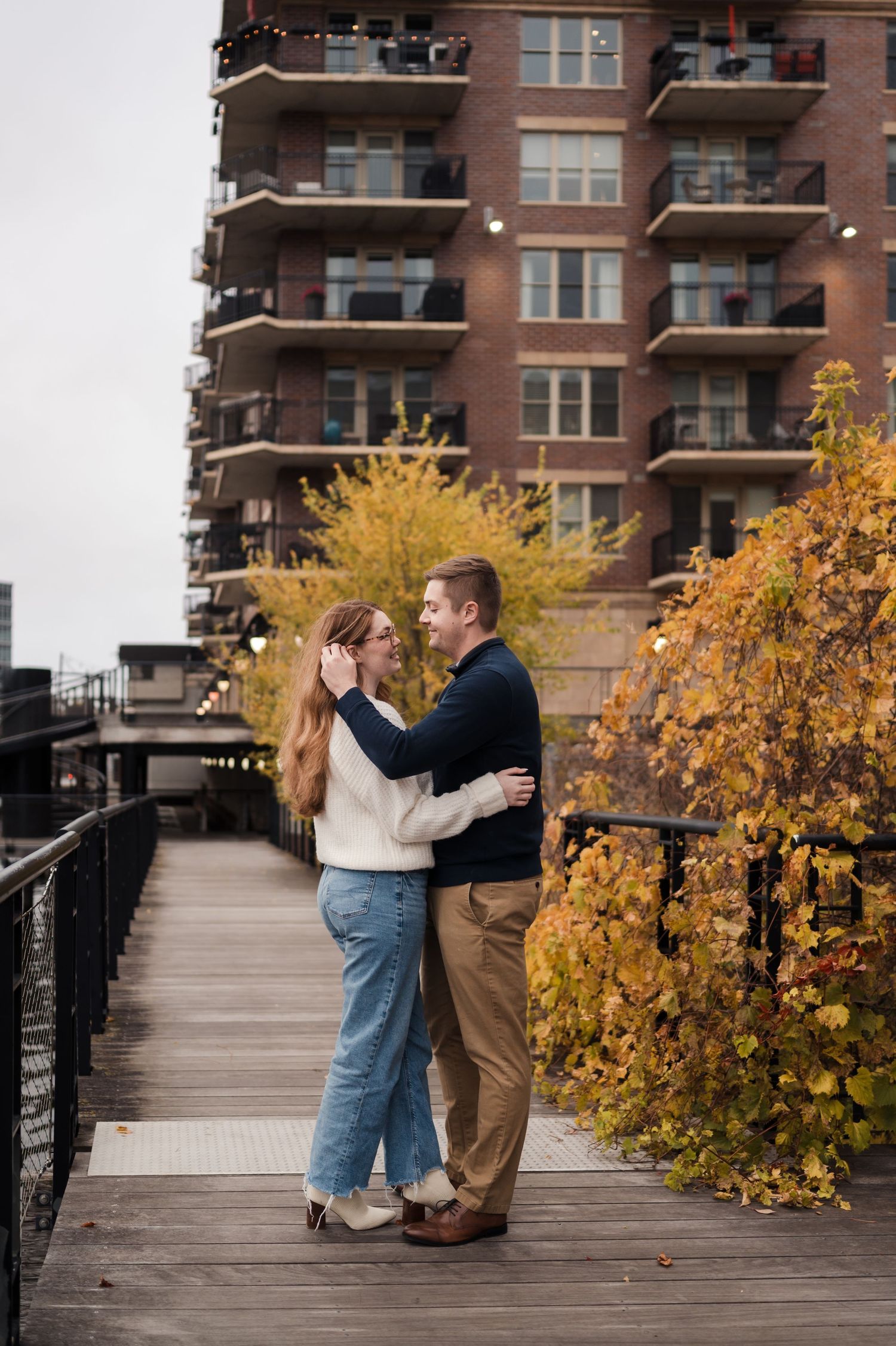 Couple embracing on a wooden boardwalk surrounded by vibrant autumn foliage with urban building backdrop.