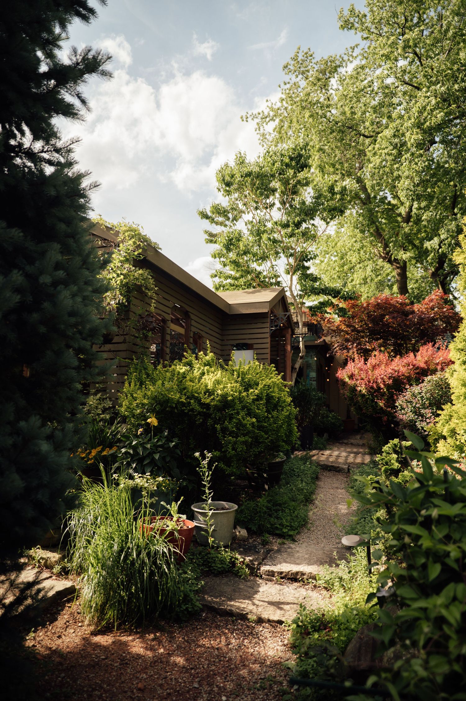 A lush garden pathway winds past a rustic wooden cabin surrounded by vibrant trees and colorful shrubs.