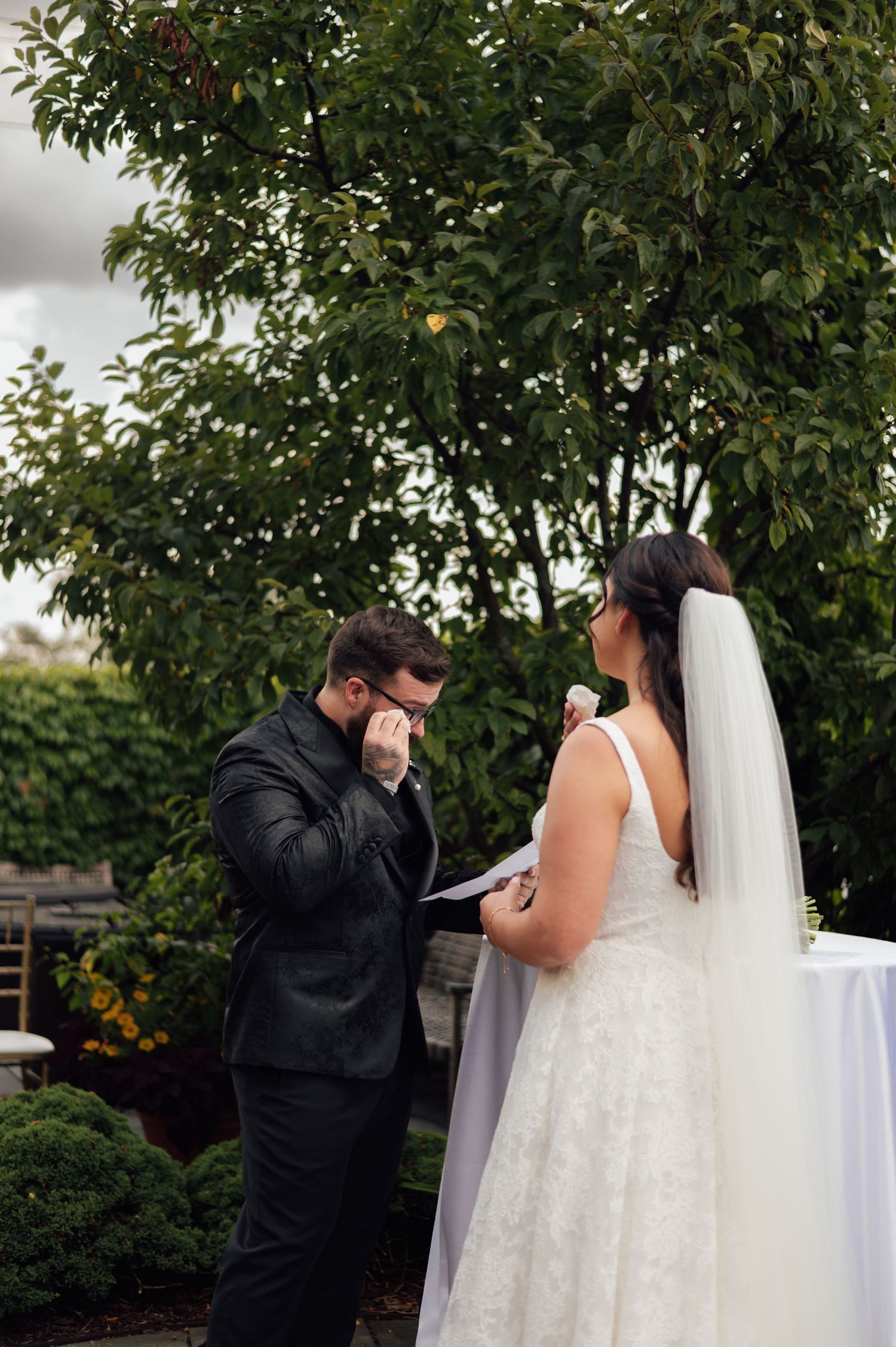 Emotional groom wiping tears during outdoor wedding ceremony while bride stands in white lace gown and veil.