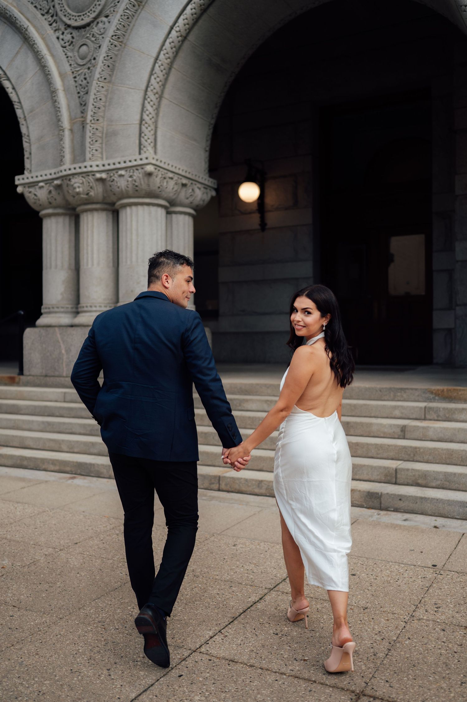 Couple holding hands walking toward ornate stone building columns, woman in white backless dress, man in navy suit.