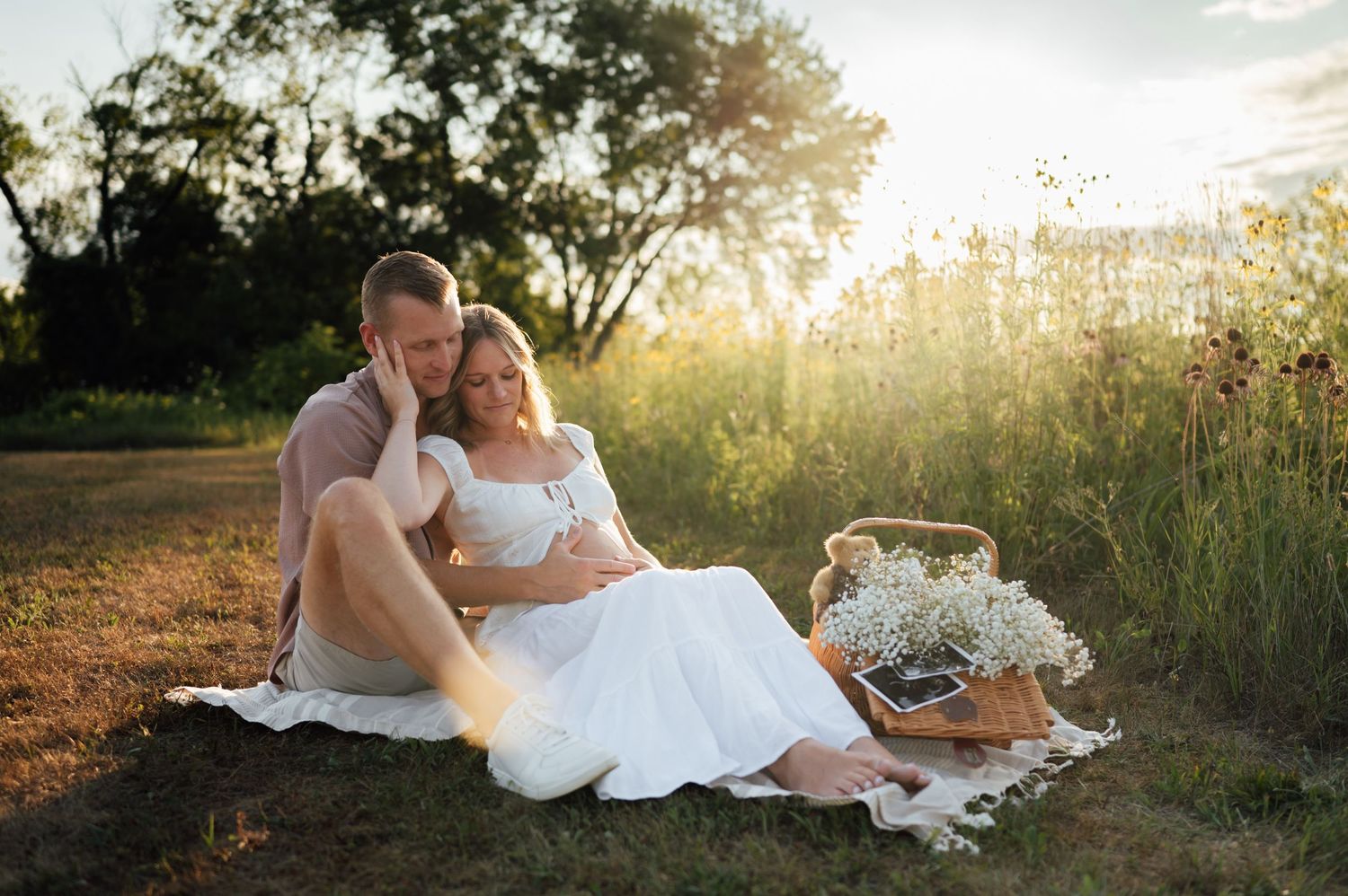 Couple sitting on blanket in golden sunset field, pregnant woman in white dress leaning on partner.