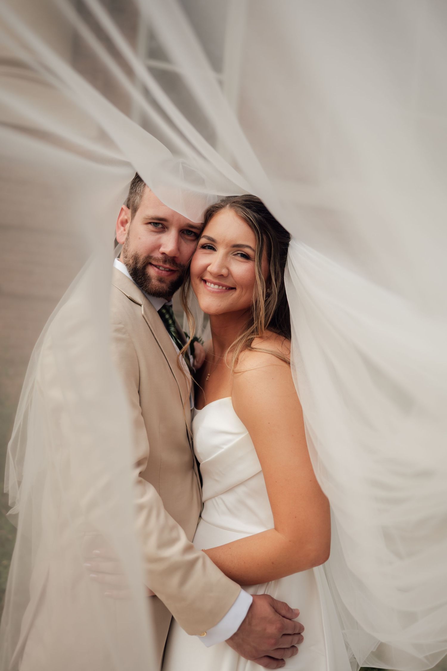 Bride and groom embracing under flowing white veil, wearing white strapless gown and tan suit, smiling at camera.