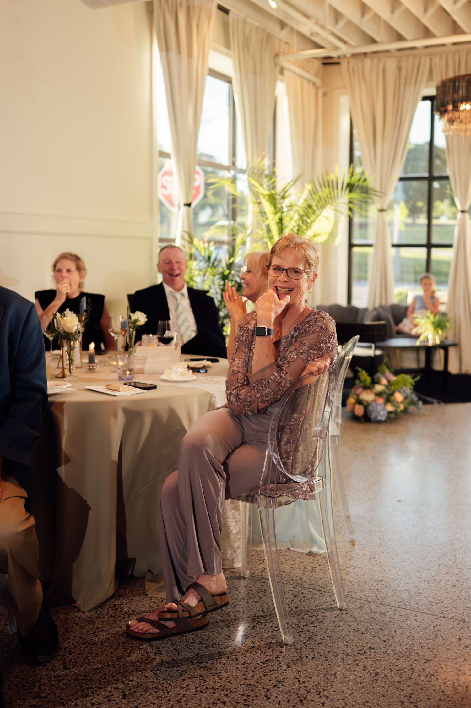 Wedding guest in grey outfit sitting on clear chair laughing during indoor reception ceremony.