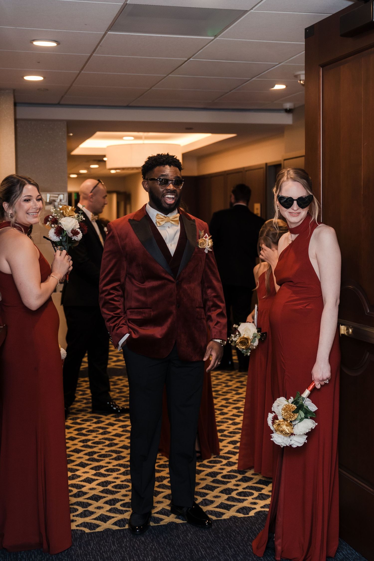 Wedding party members in burgundy dresses and black suits gather in a carpeted hallway with warm lighting.