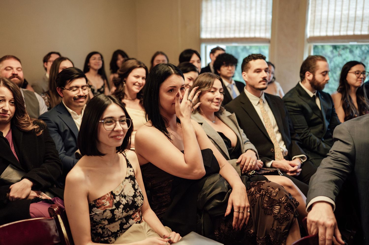 Guests in formal attire smile and react emotionally during an indoor wedding ceremony seated in rows.